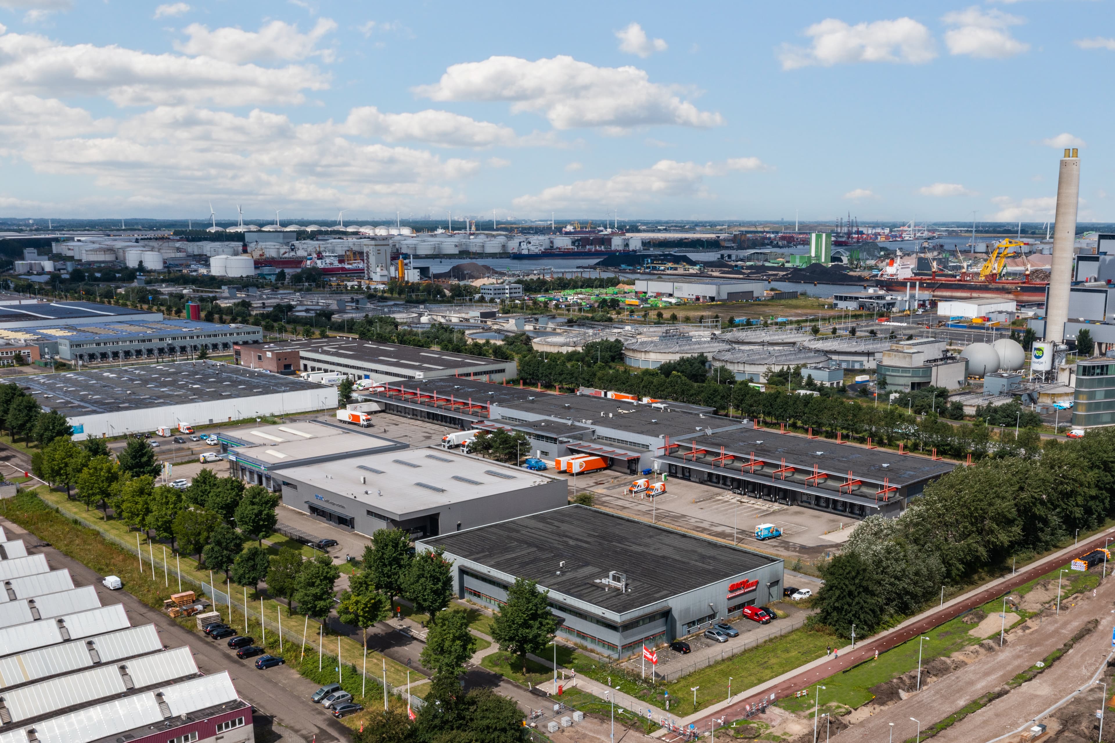 Aerial view of an industrial area with factories, warehouses, roads, and parked vehicles under a partly cloudy sky.