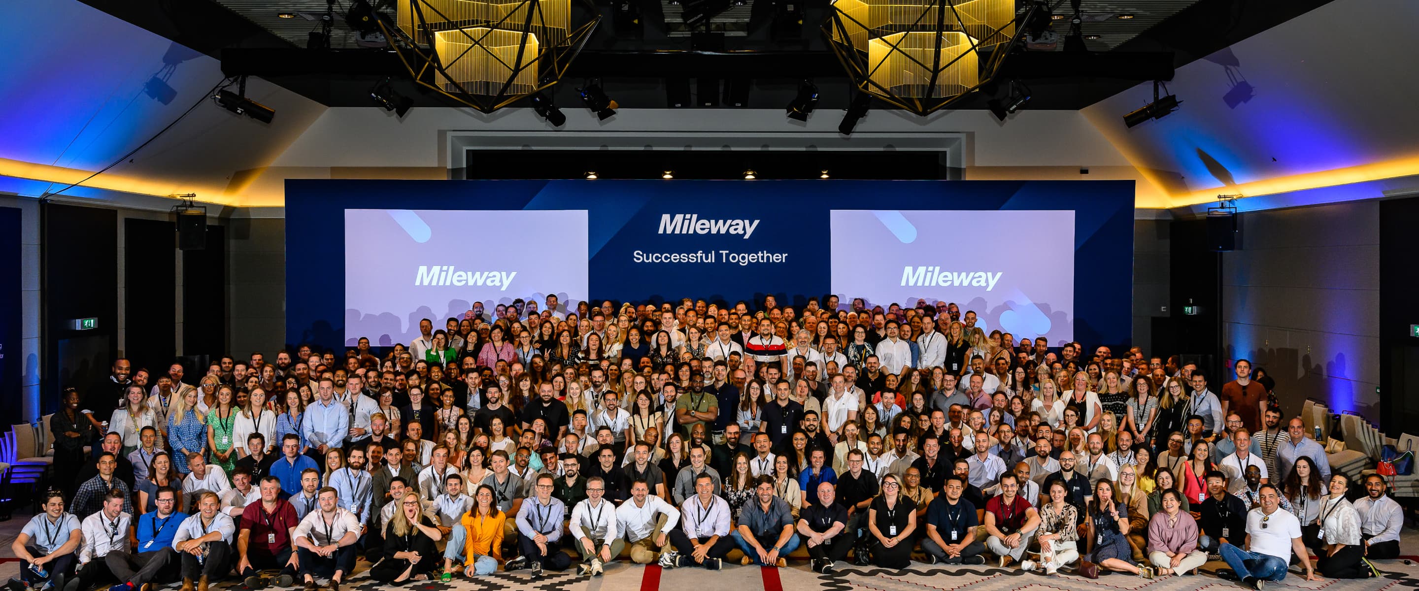 A large group of people pose together on a stage in front of two screens displaying Mileway and Successful Together.
