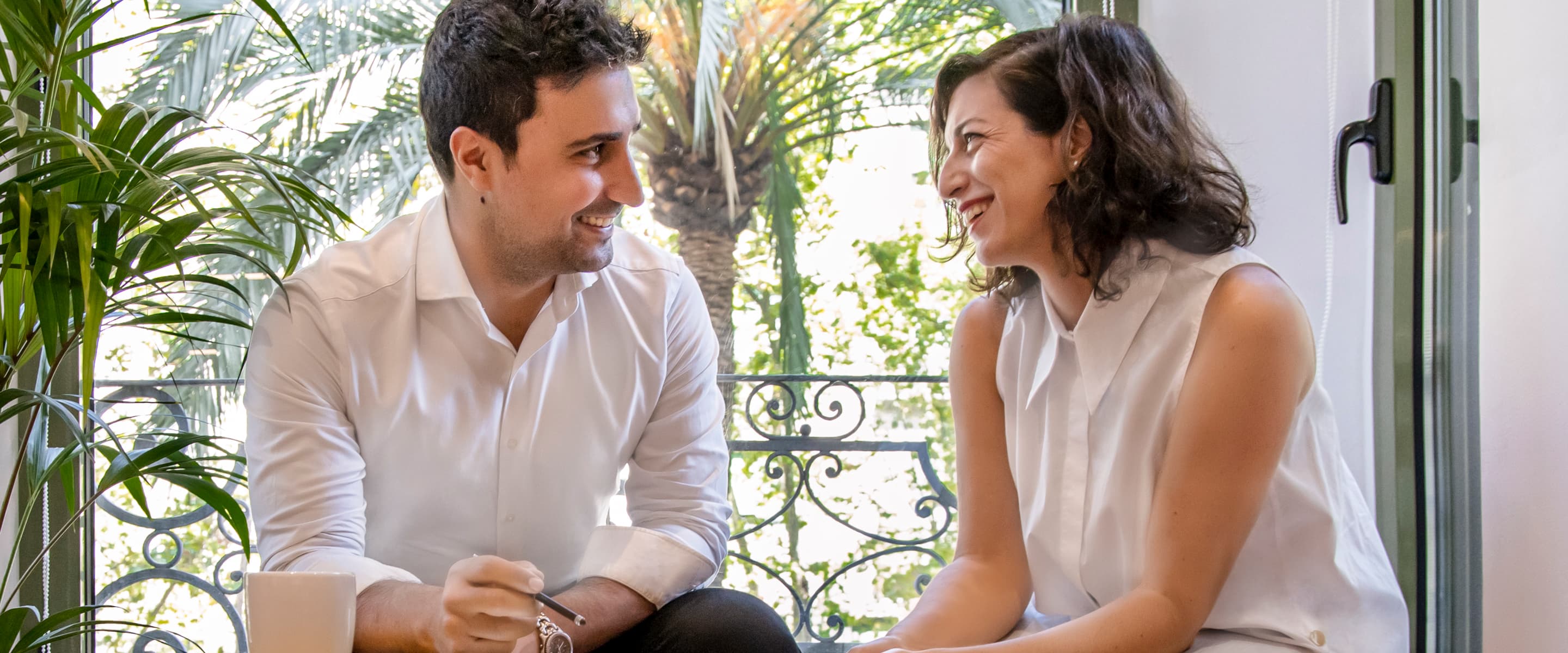 A man and a woman sit and smile at each other in a bright room with plants and a window in the background.
