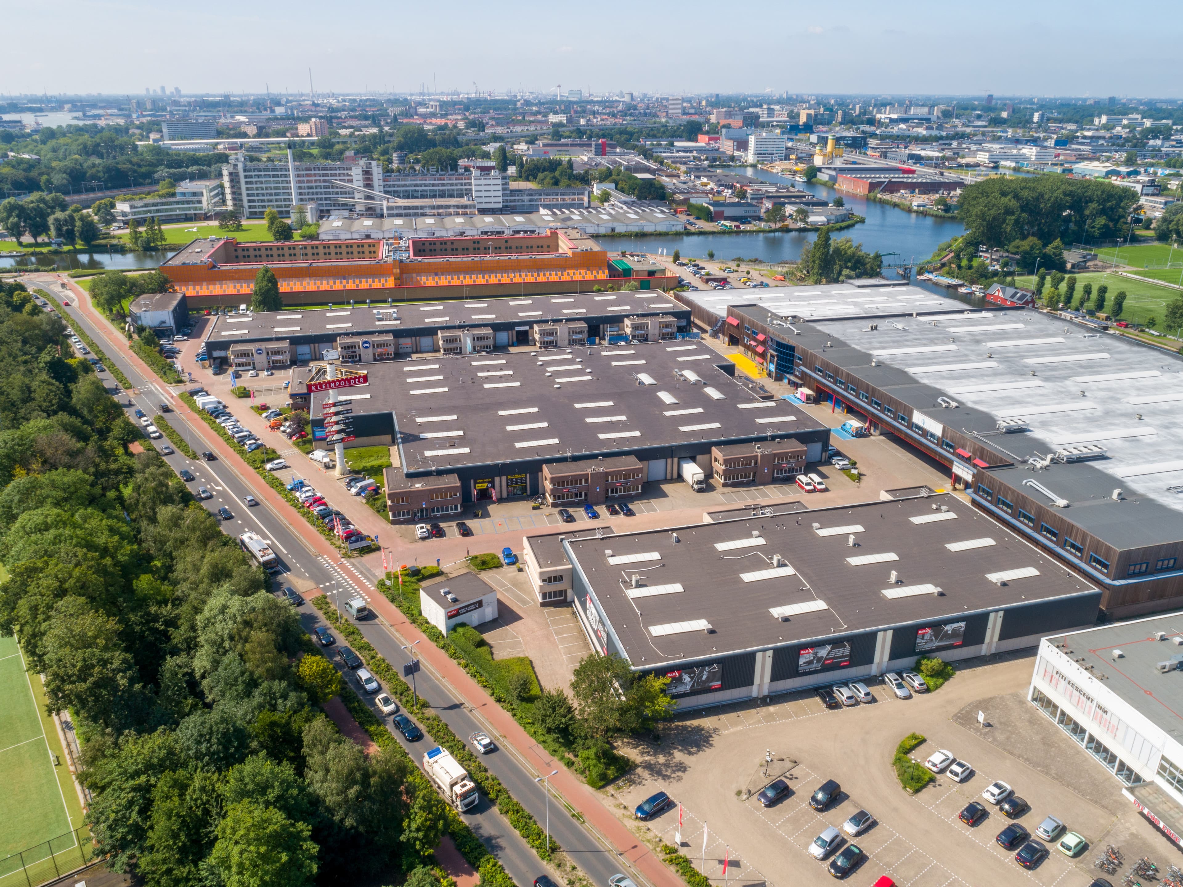 Aerial view of an industrial park with large warehouse buildings, roads, and surrounding greenery. The urban landscape extends into the distance under a clear blue sky.