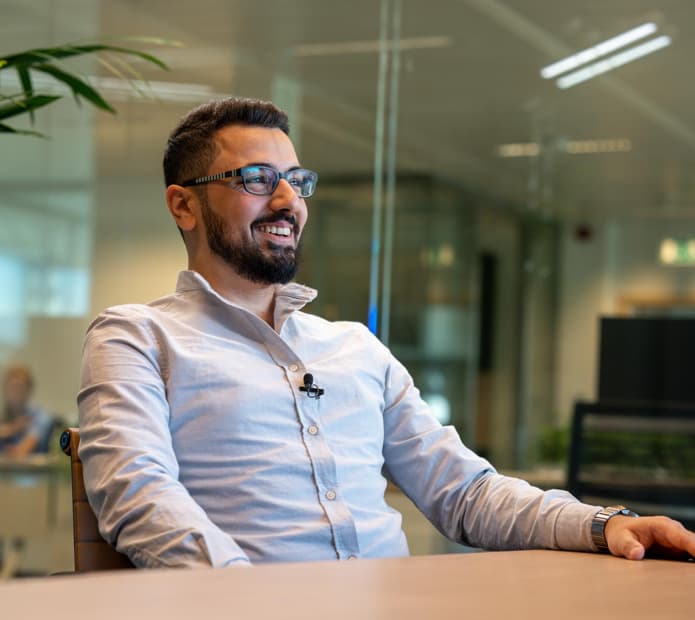Man with glasses and beard sitting at a table, smiling, indoors with a blurred office background.