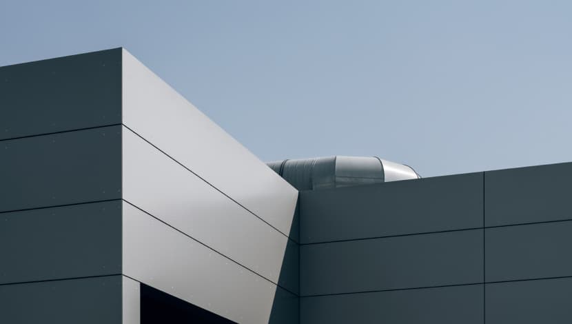 Modern building corner with overlapping metal panels and a visible air duct under a clear sky.