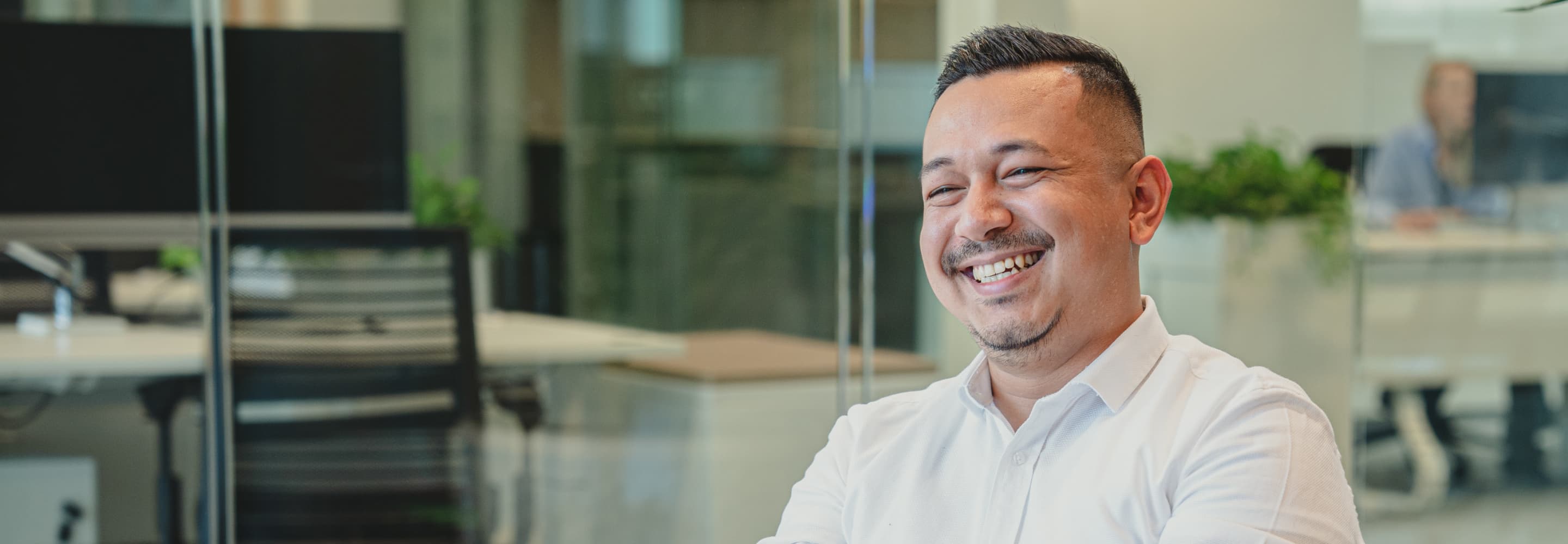 A man in a white shirt is smiling, seated in an office environment with glass partitions and desk chairs in the background.