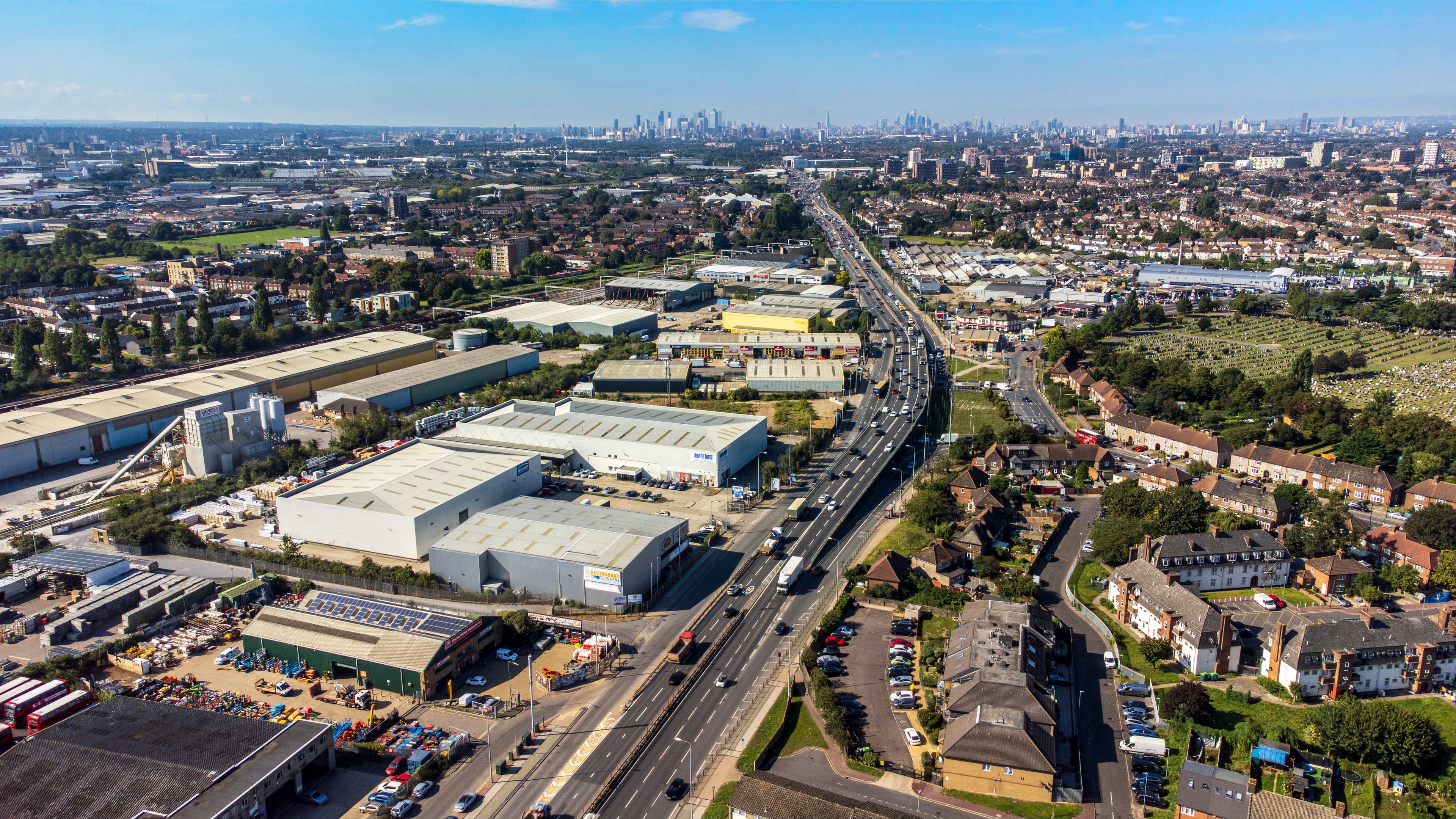 A modern logistics facility with a large, clean, white exterior, multiple loading bays, and a spacious parking area, surrounded by greenery and under a partly cloudy sky