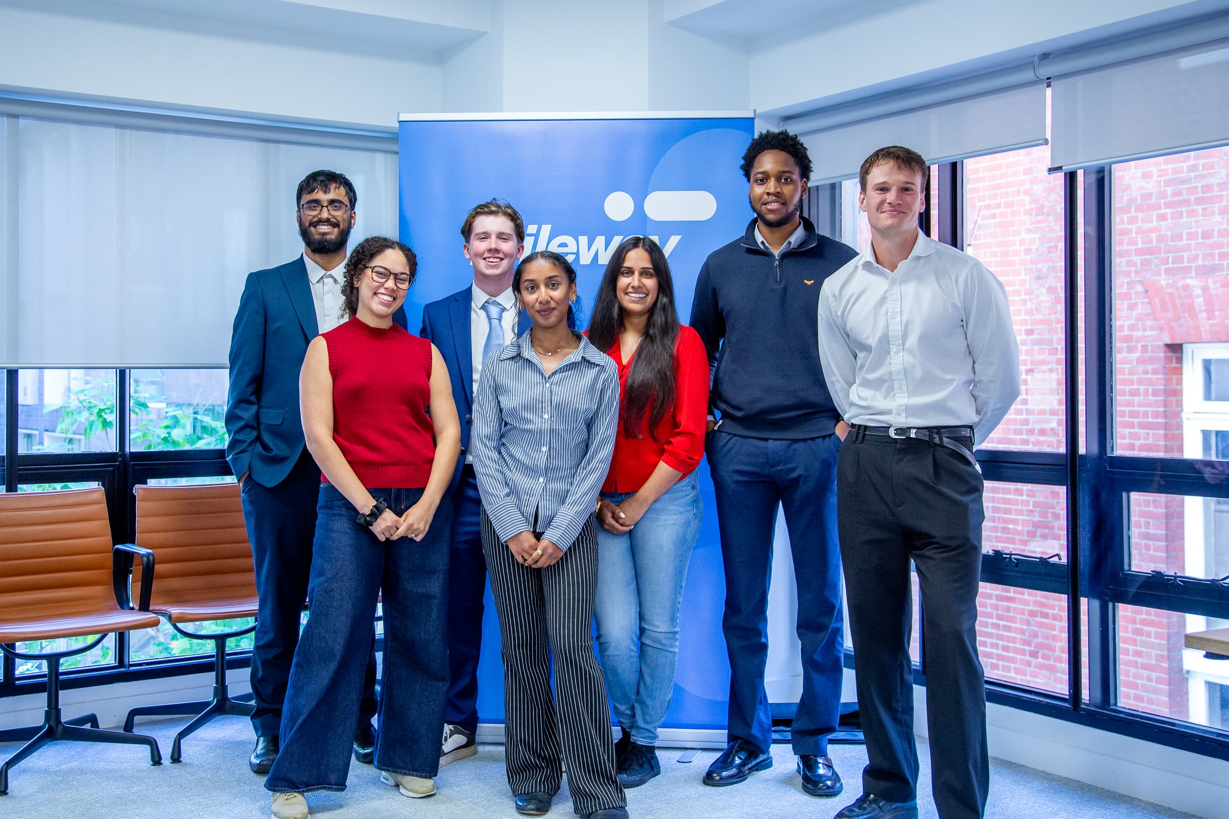 Seven people stand and pose for a group photo in an office setting, in front of a blue banner with the Mileway logo. Some are smiling; all are dressed in business or business casual attire.