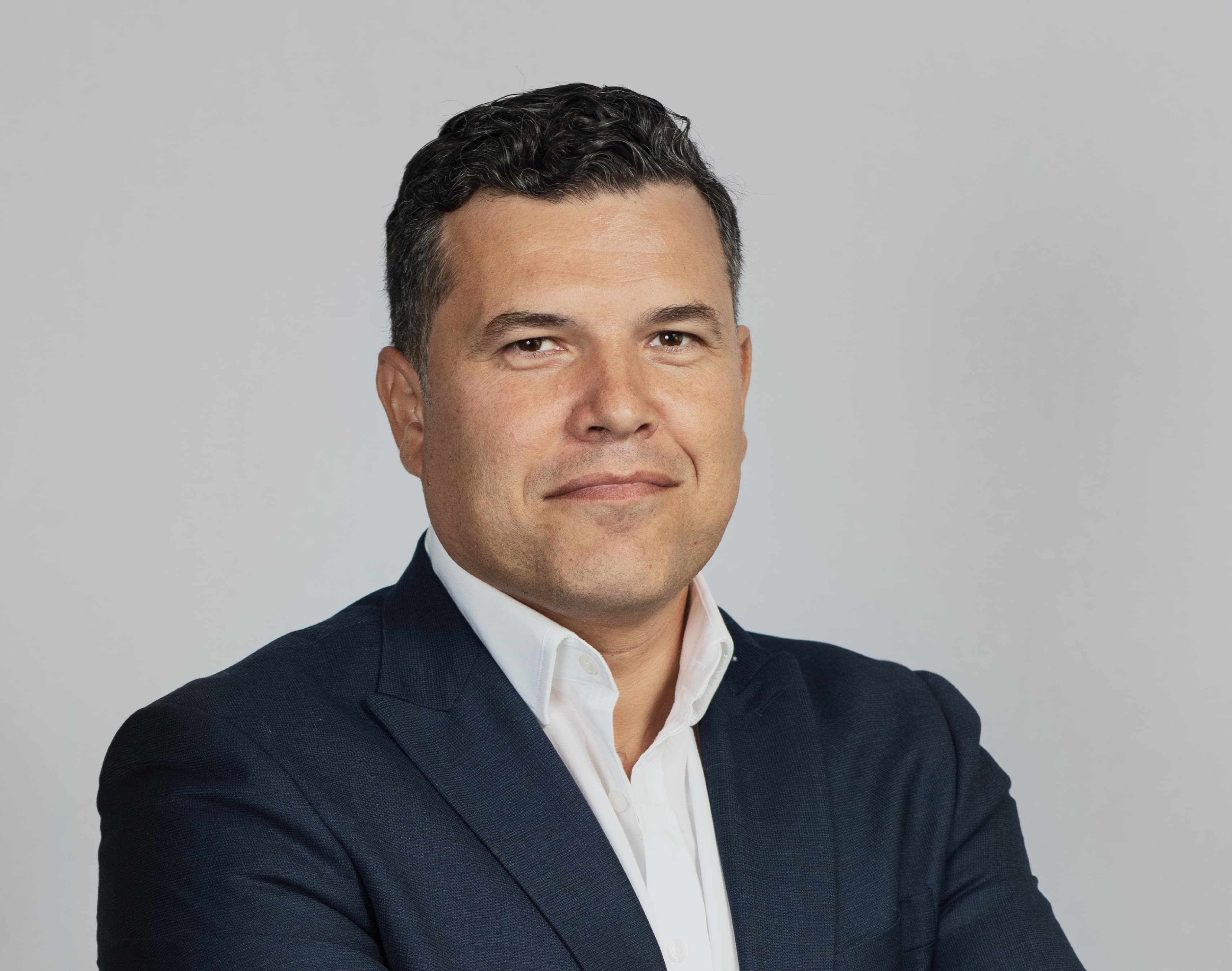 A man in a dark suit and white shirt stands against a plain gray background with a neutral expression.
