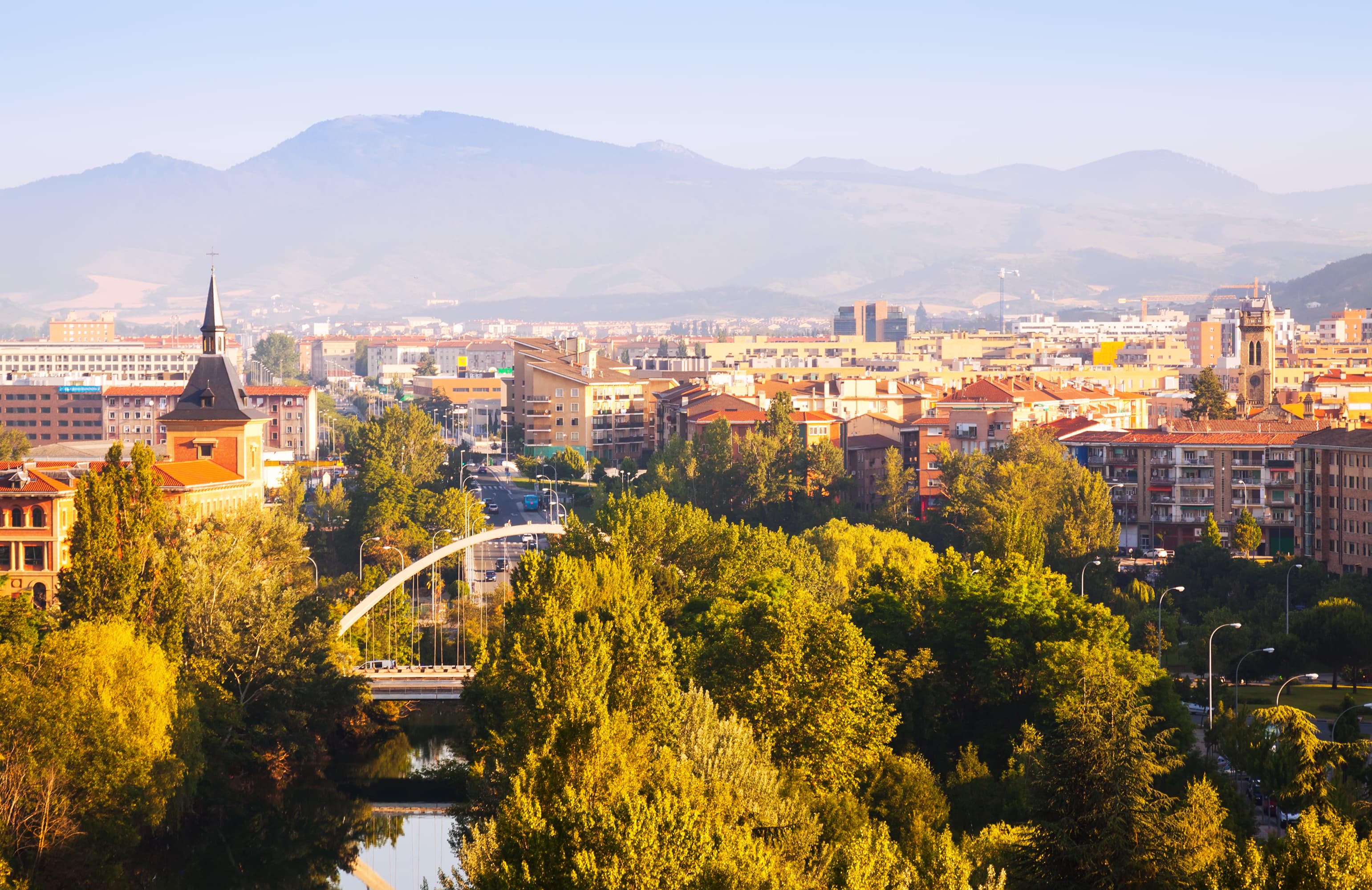 Aerial view of a cityscape with dense greenery, modern and historic buildings, and mountains in the background under a clear blue sky.