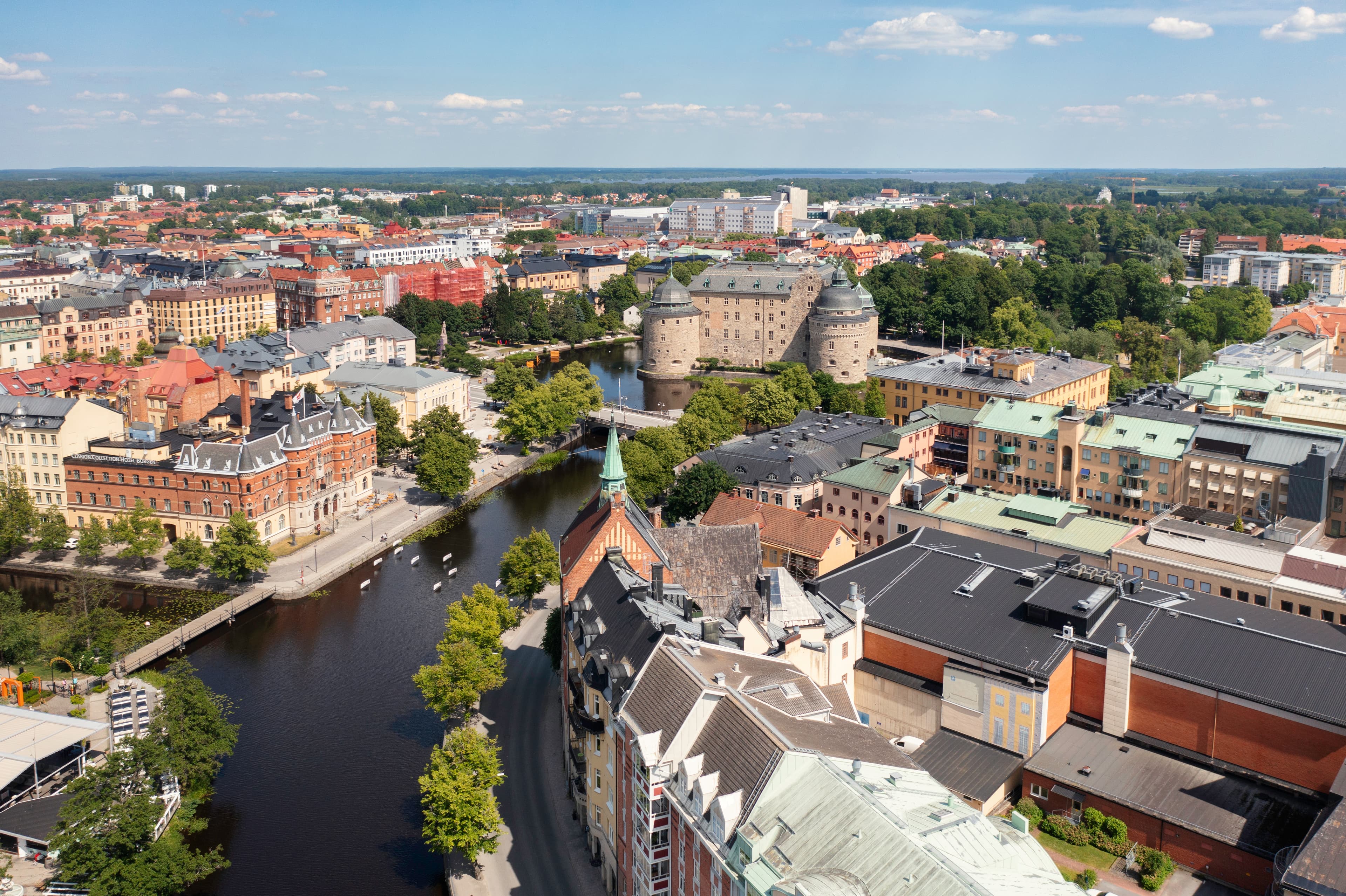 Aerial view of a cityscape featuring a river, a castle with round towers, and surrounding buildings under a partly cloudy sky.