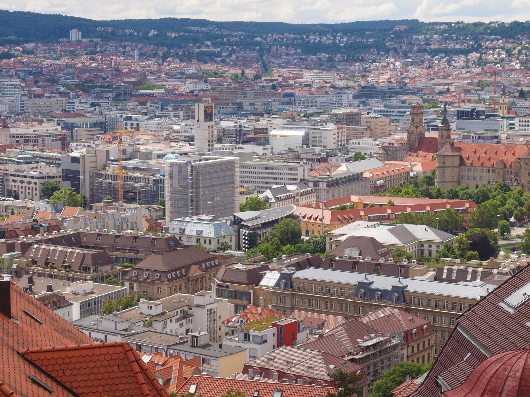 Aerial view of a cityscape with a mix of modern and historic buildings, green spaces, and hills in the background under a partly cloudy sky.