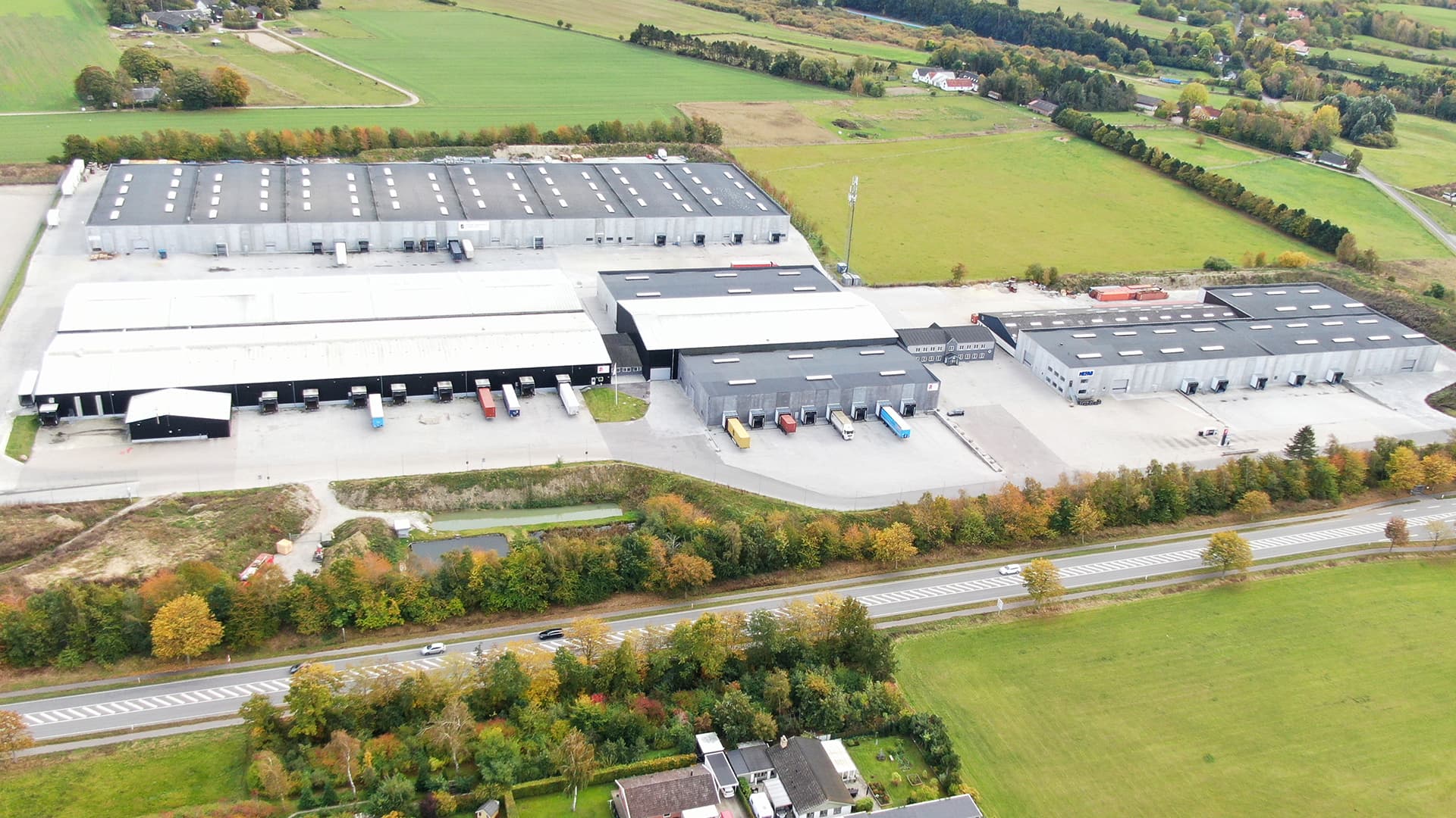 Aerial view of a large industrial complex with warehouses, parking areas, and trucks. Surrounded by green fields and a road in the foreground.
