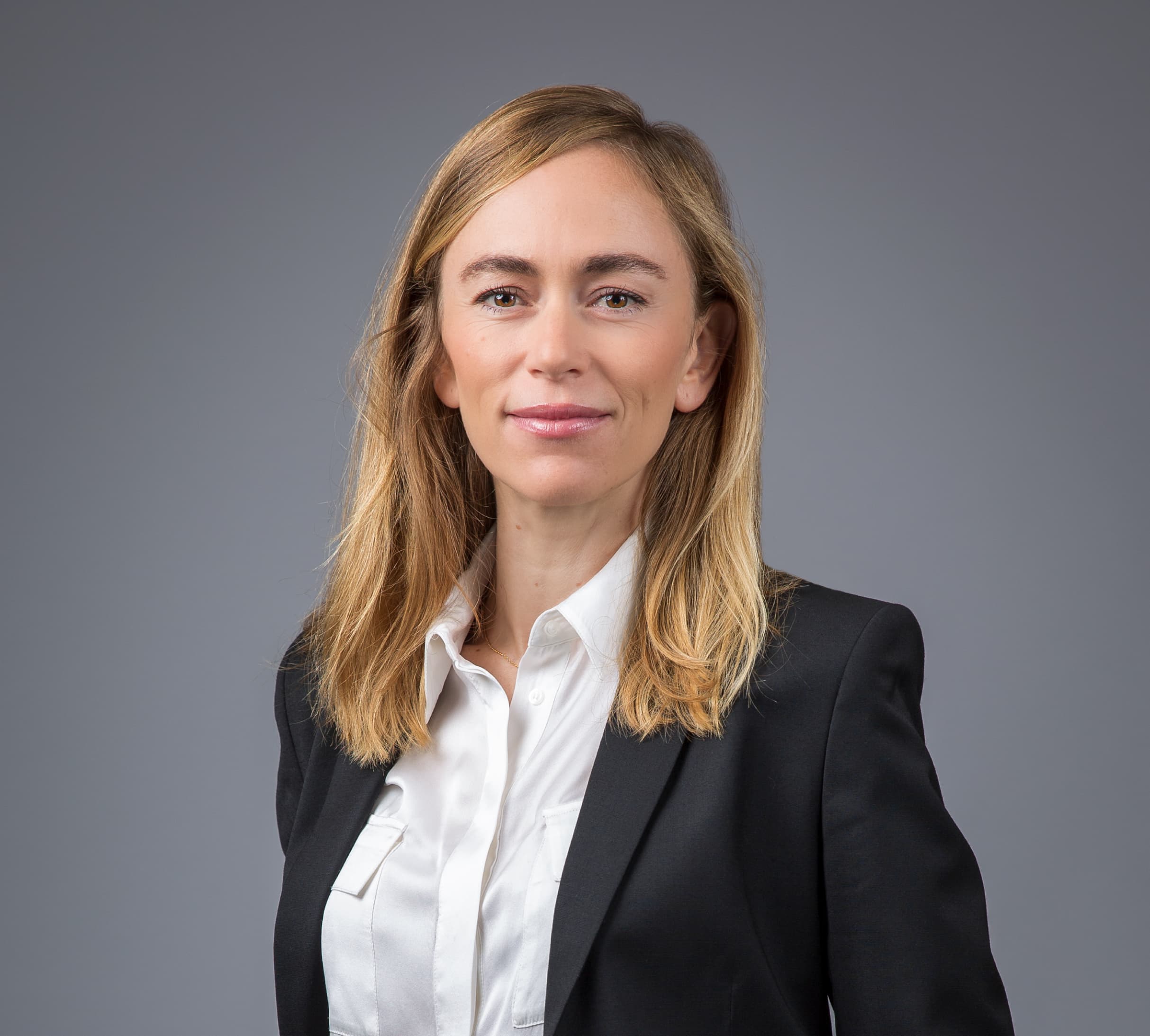 A woman with shoulder-length blond hair is wearing a white shirt and black blazer, posing against a gray background.