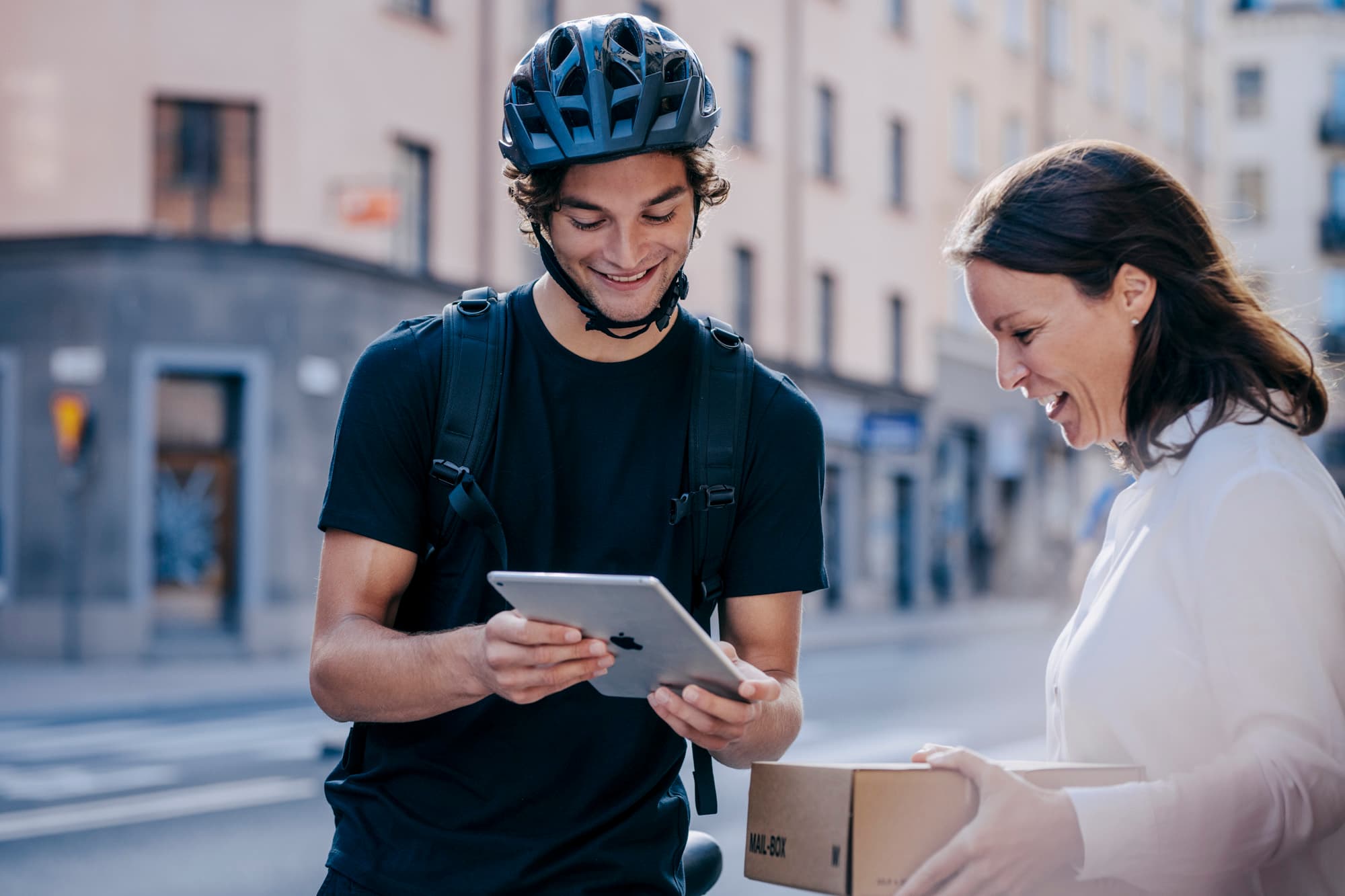 A cyclist wearing a helmet hands a tablet to a woman holding a box on a city street.