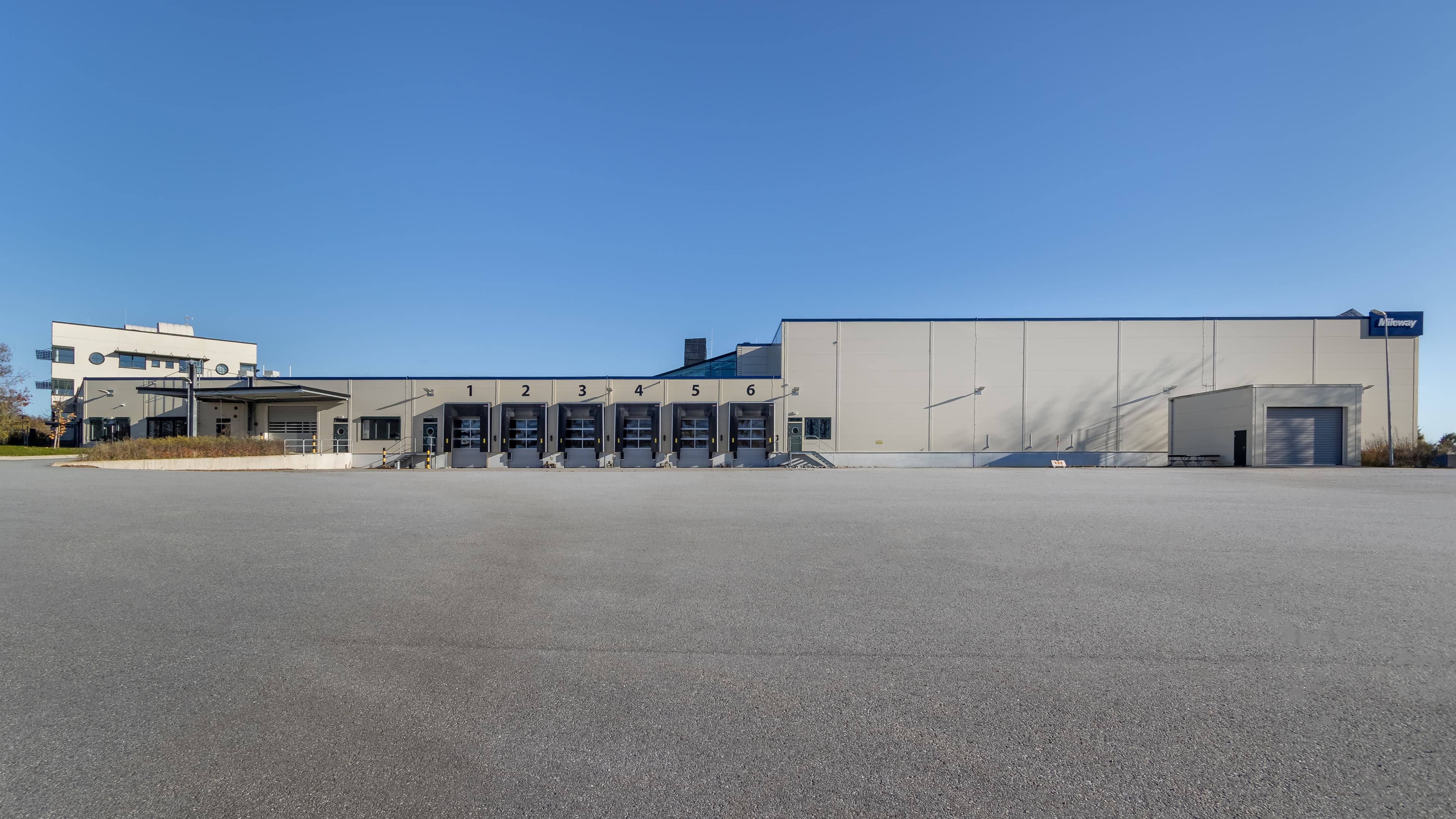 A large industrial warehouse with loading docks numbered 1 to 6, situated on a vast concrete lot under a clear blue sky.