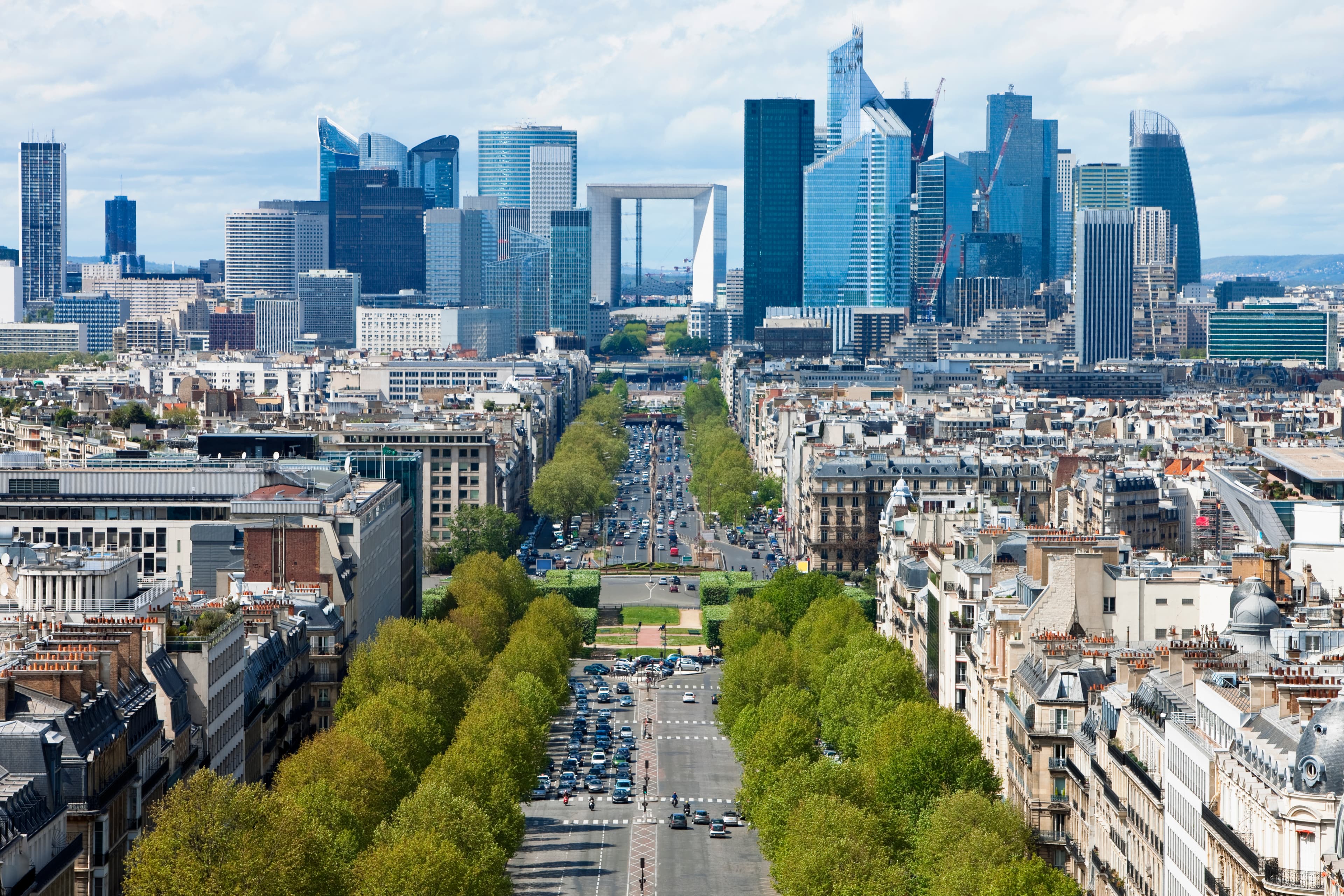 Aerial view of a cityscape featuring a wide boulevard lined with trees and leading to a modern skyscraper district in the distance.