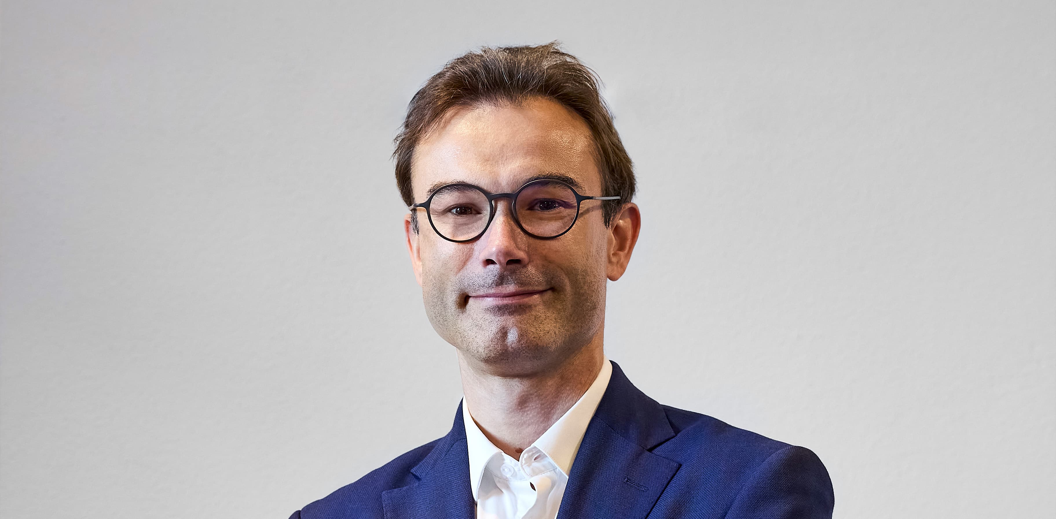 A man in a blue suit and glasses stands against a plain background, smiling slightly.