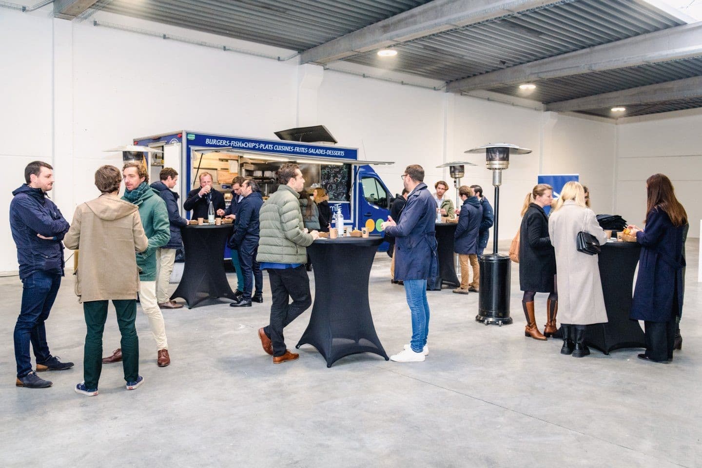 People gather around black tables near a blue food truck in a spacious indoor setting.