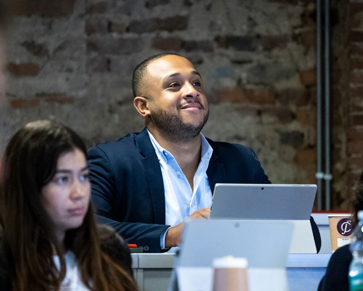 A man in a suit sits at a desk, smiling and looking forward. A woman is seated in front, focusing on something outside the frame.