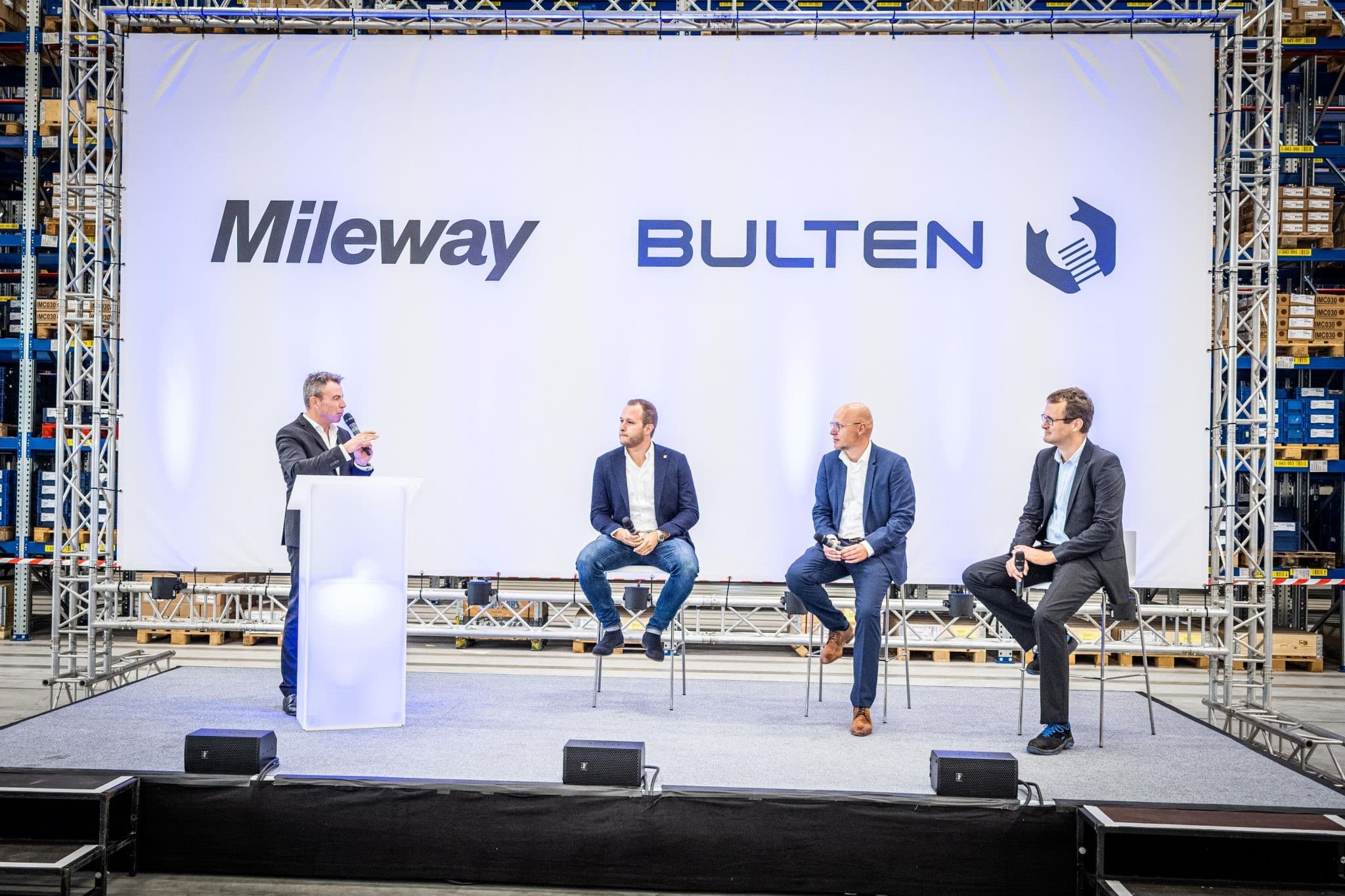 Four men in business attire sit on stools on a stage with Mileway and Bulten logos on the backdrop. One man is speaking at a podium.