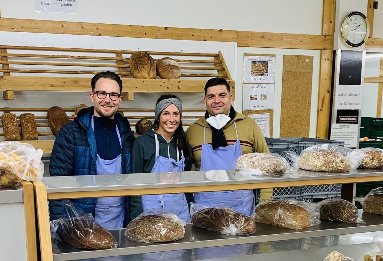 Three people wearing aprons stand behind a display of various breads in a bakery setting.