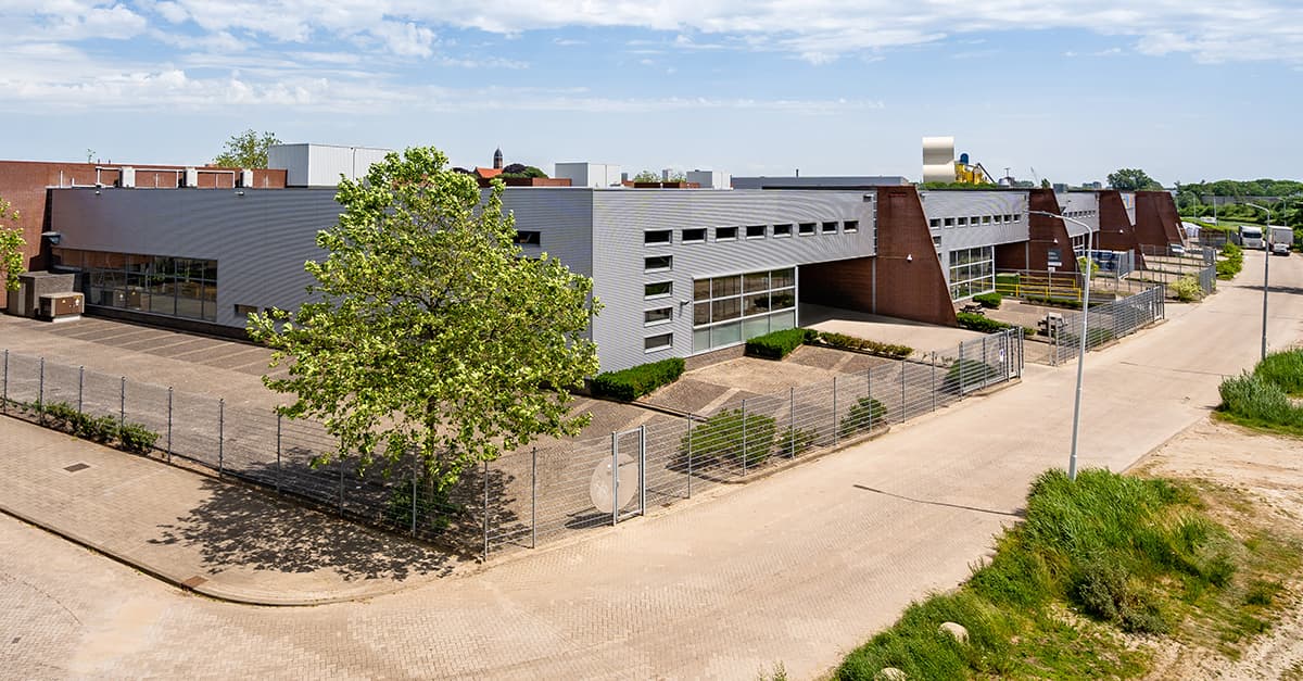 Modern industrial building with a metal exterior, surrounded by a fence and trees, located on a paved road under a partly cloudy sky.