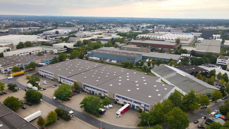 Aerial view of an industrial park with large rectangular buildings, surrounding roadways, and parked vehicles. Urban landscape and cloudy sky in the background.