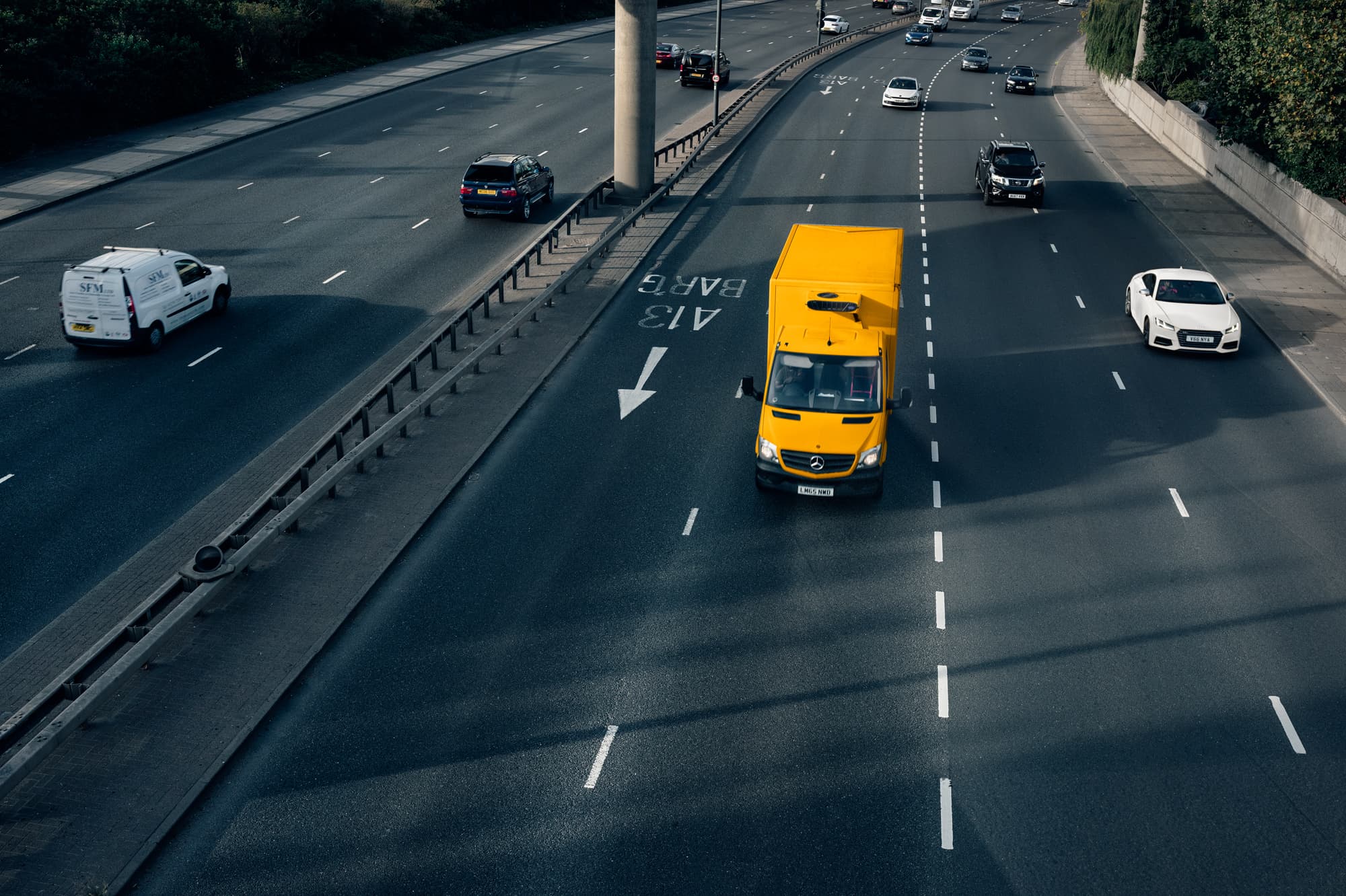 A yellow delivery van drives on a multilane highway alongside other vehicles, surrounded by trees and barriers.