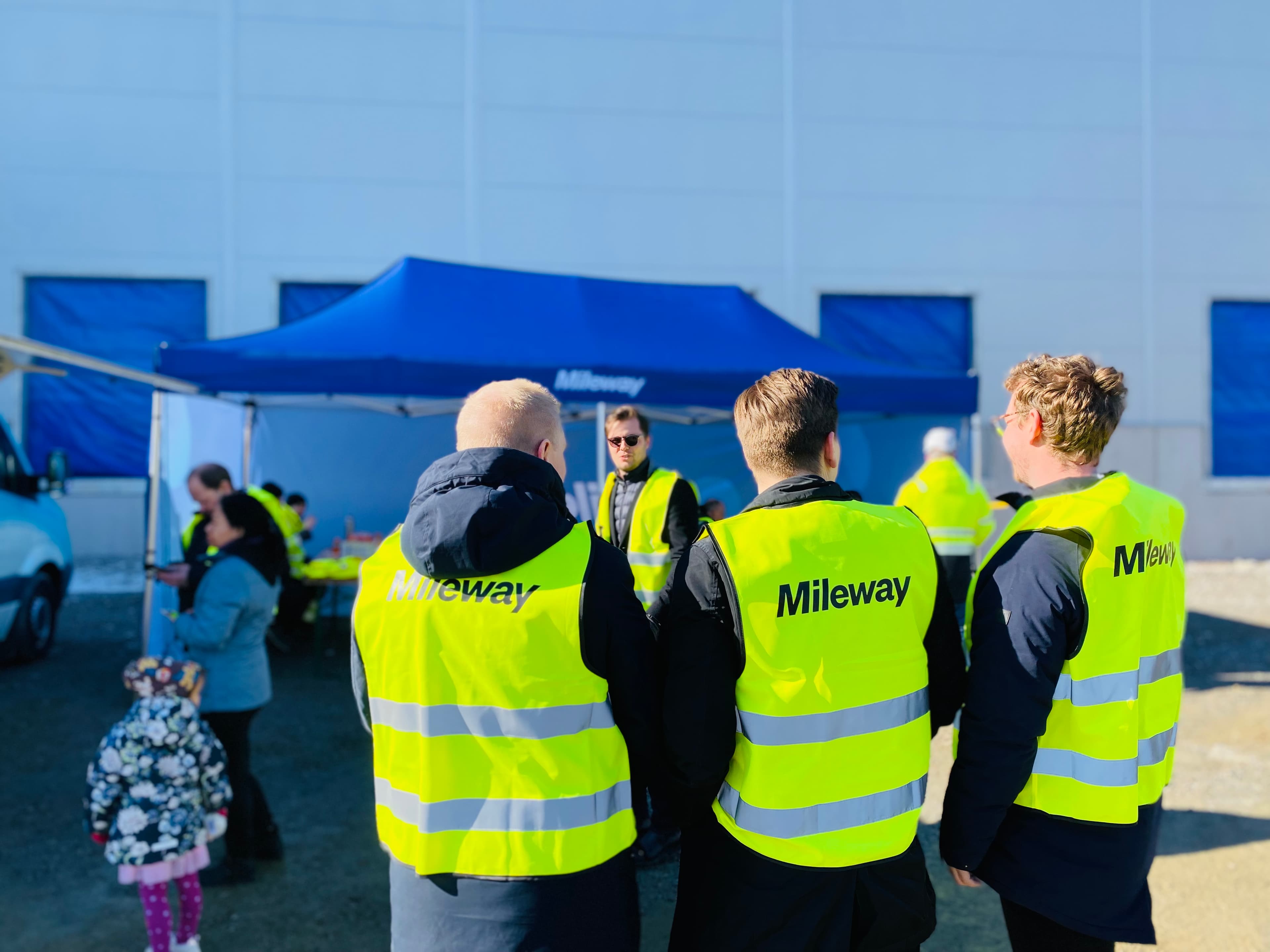 Several people in yellow vests with Mileway written on them stand near a blue tent outside. More people are in the background along with a child in a floral jacket.