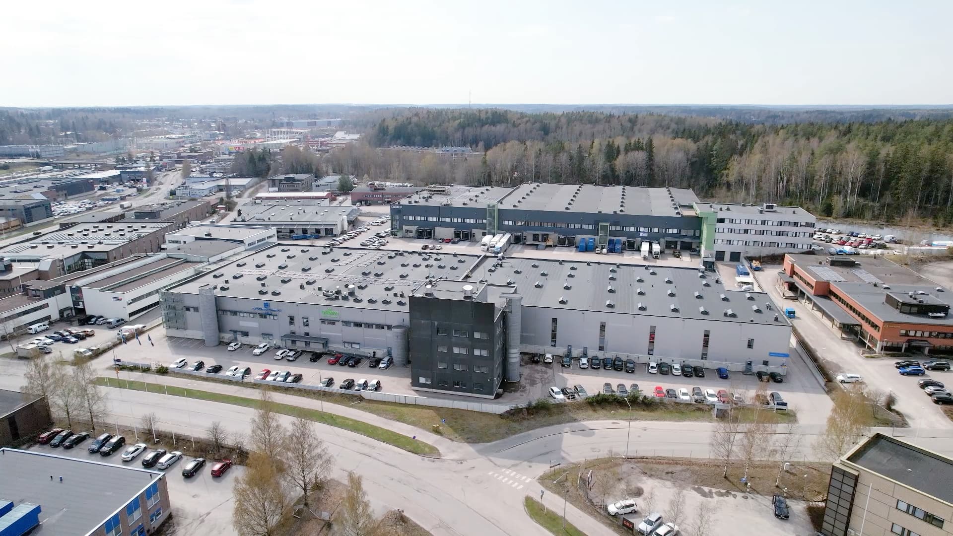Aerial view of an industrial complex with multiple buildings and a parking lot filled with cars, surrounded by trees and nearby roads.