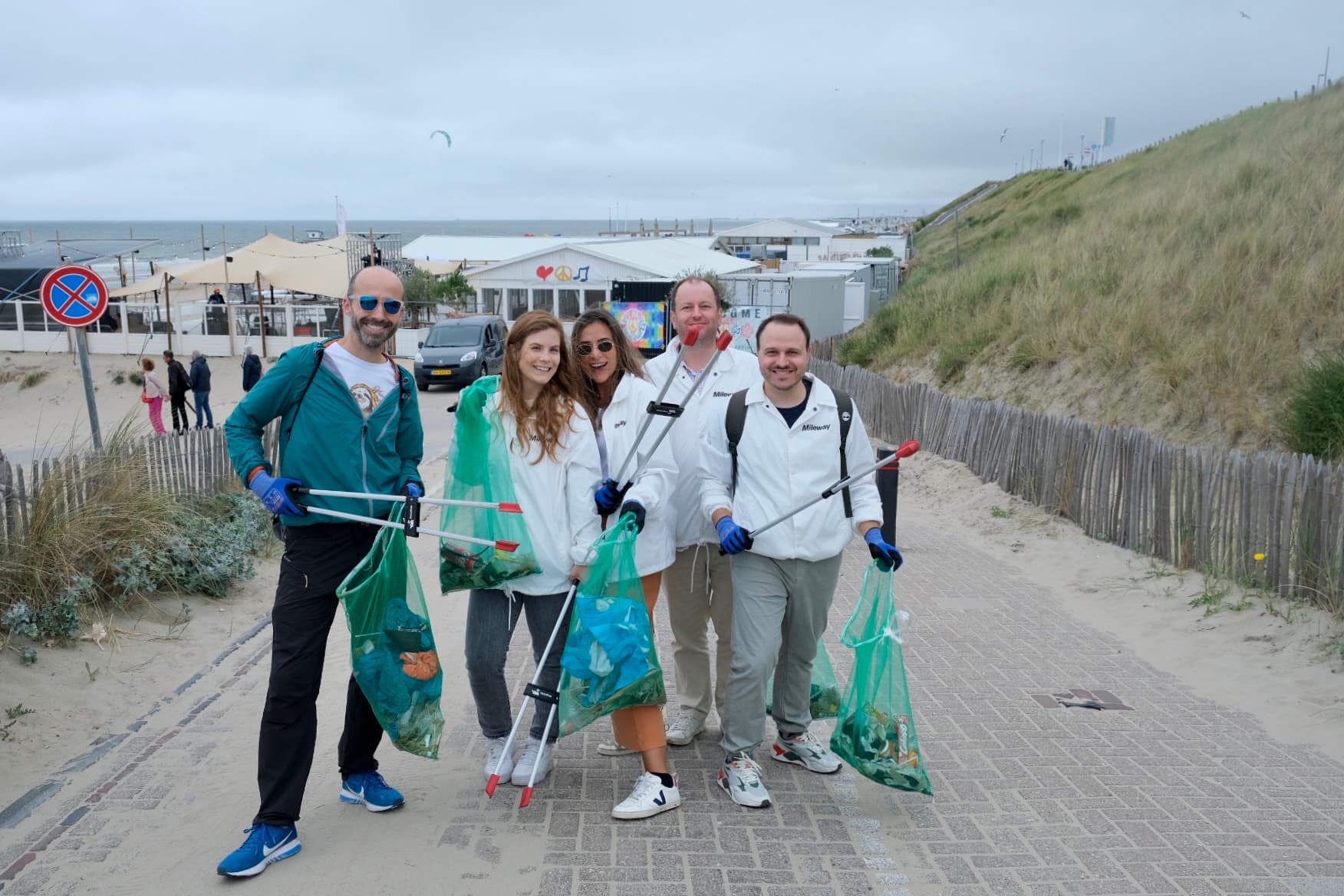 A group of five people holding trash bags and litter pickers clean up a beach path. They are smiling and dressed in casual clothes. Sand dunes and beach umbrellas are in the background.