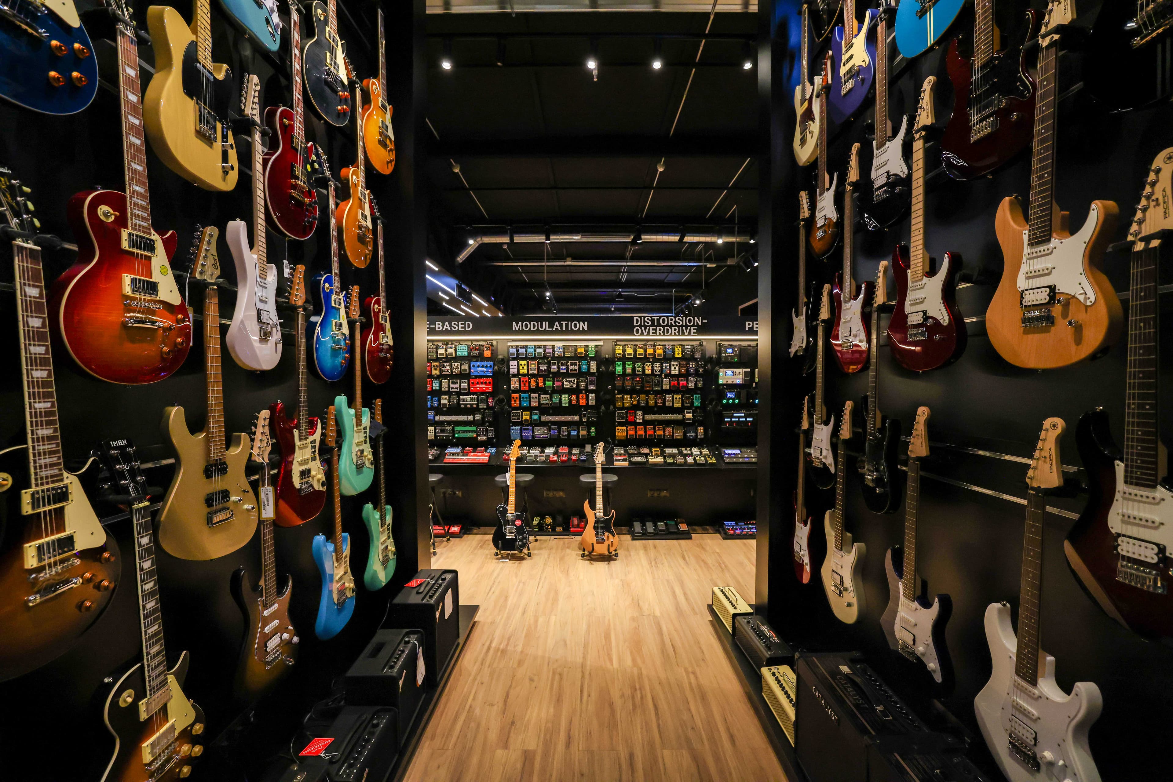 A music store aisle with electric guitars on both sides, showcasing various models and colors. Floor displays feature amplifiers.