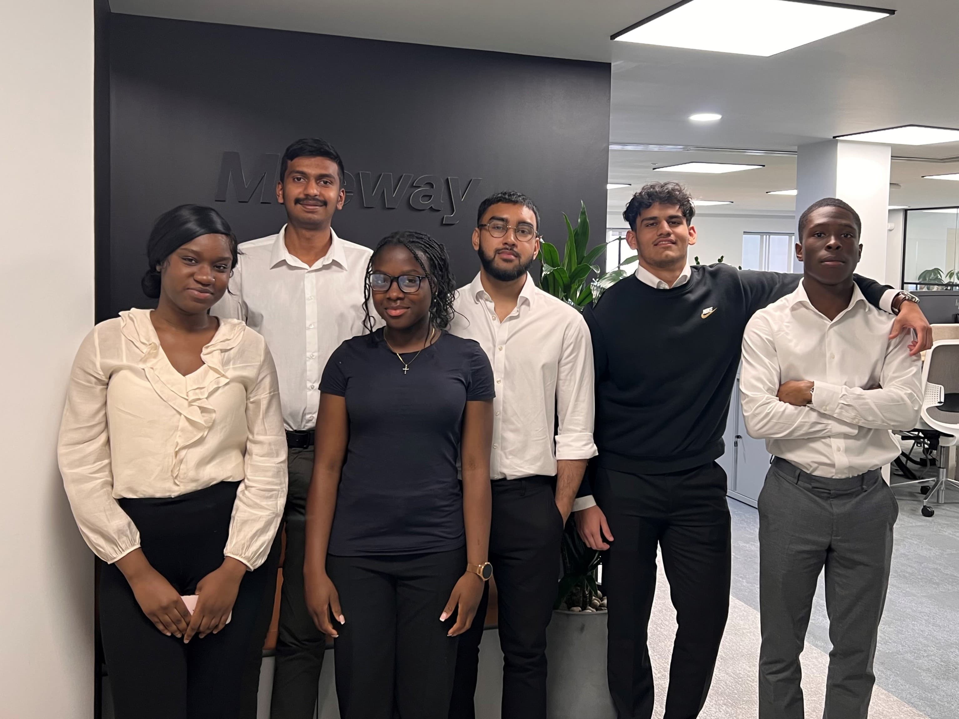 Six people standing indoors in a row, wearing business casual attire, against a black wall with the word Moway partially visible.