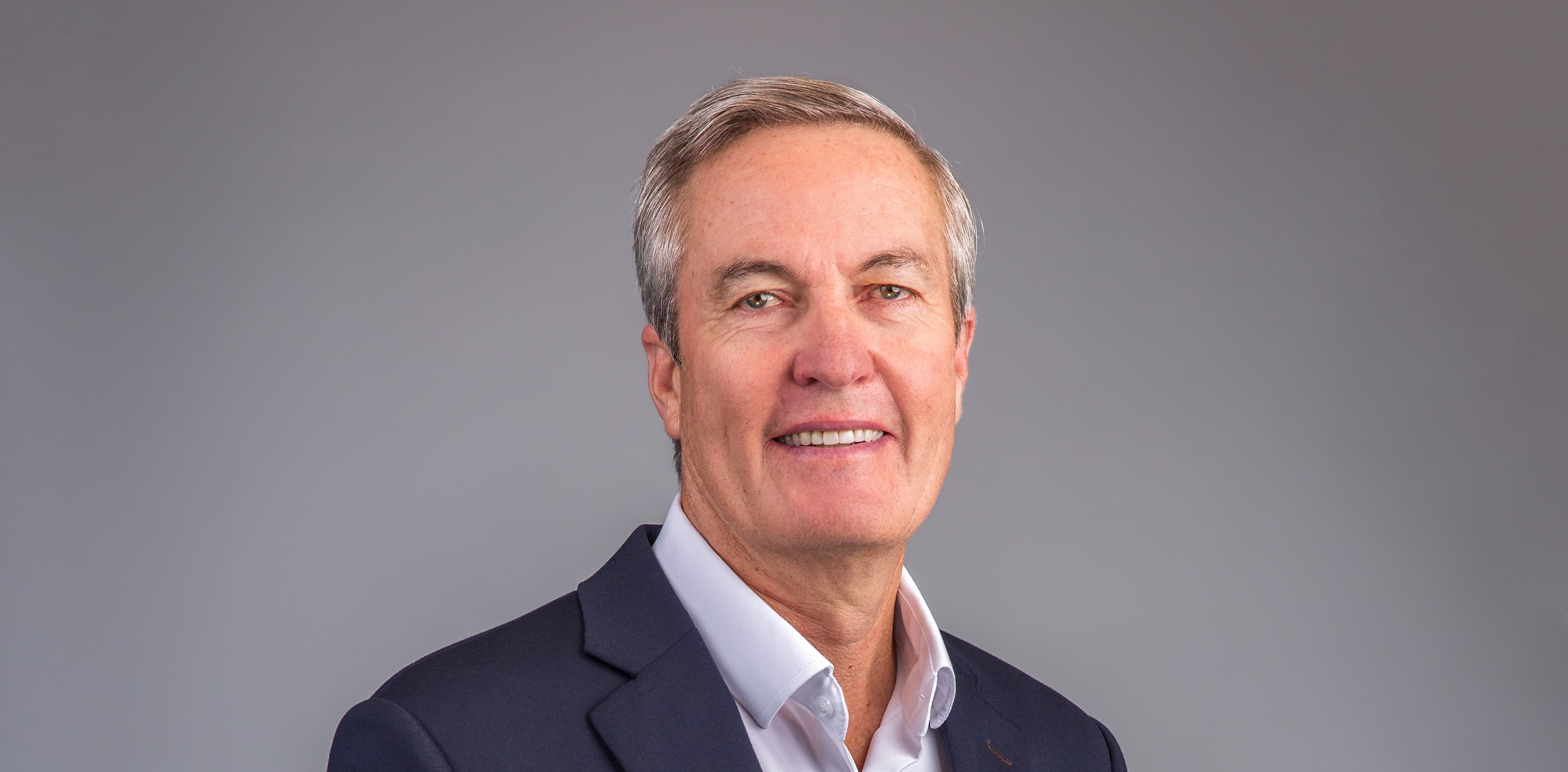A middle-aged man in a suit and white shirt poses against a plain gray background.