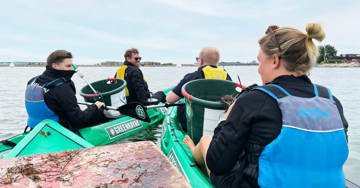 Four people in life vests collect litter in green kayaks on a calm body of water.
