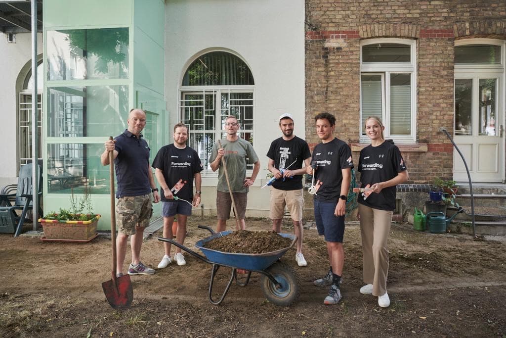 A group of six people stand outside a building. They are holding garden tools, and a wheelbarrow filled with soil is in front of them.
