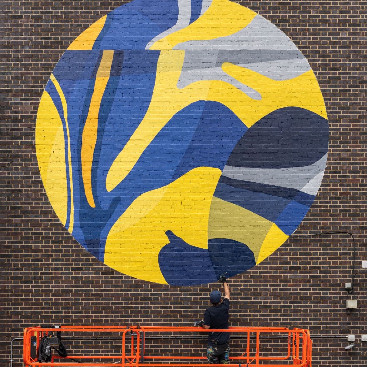 Artist on a lift paints a large abstract mural with blue, yellow, and gray shapes on a brick wall.