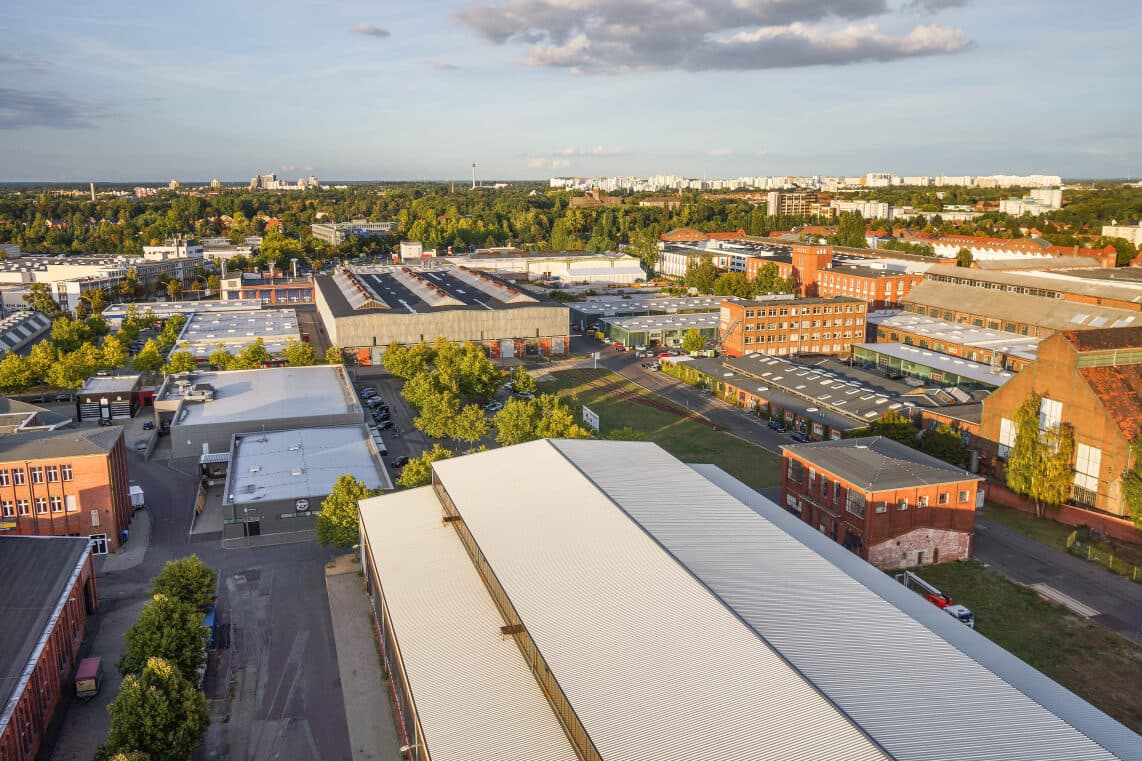 Aerial view of an industrial area with warehouses, buildings, and tree-lined streets under a partly cloudy sky. A cityscape is visible in the distance.