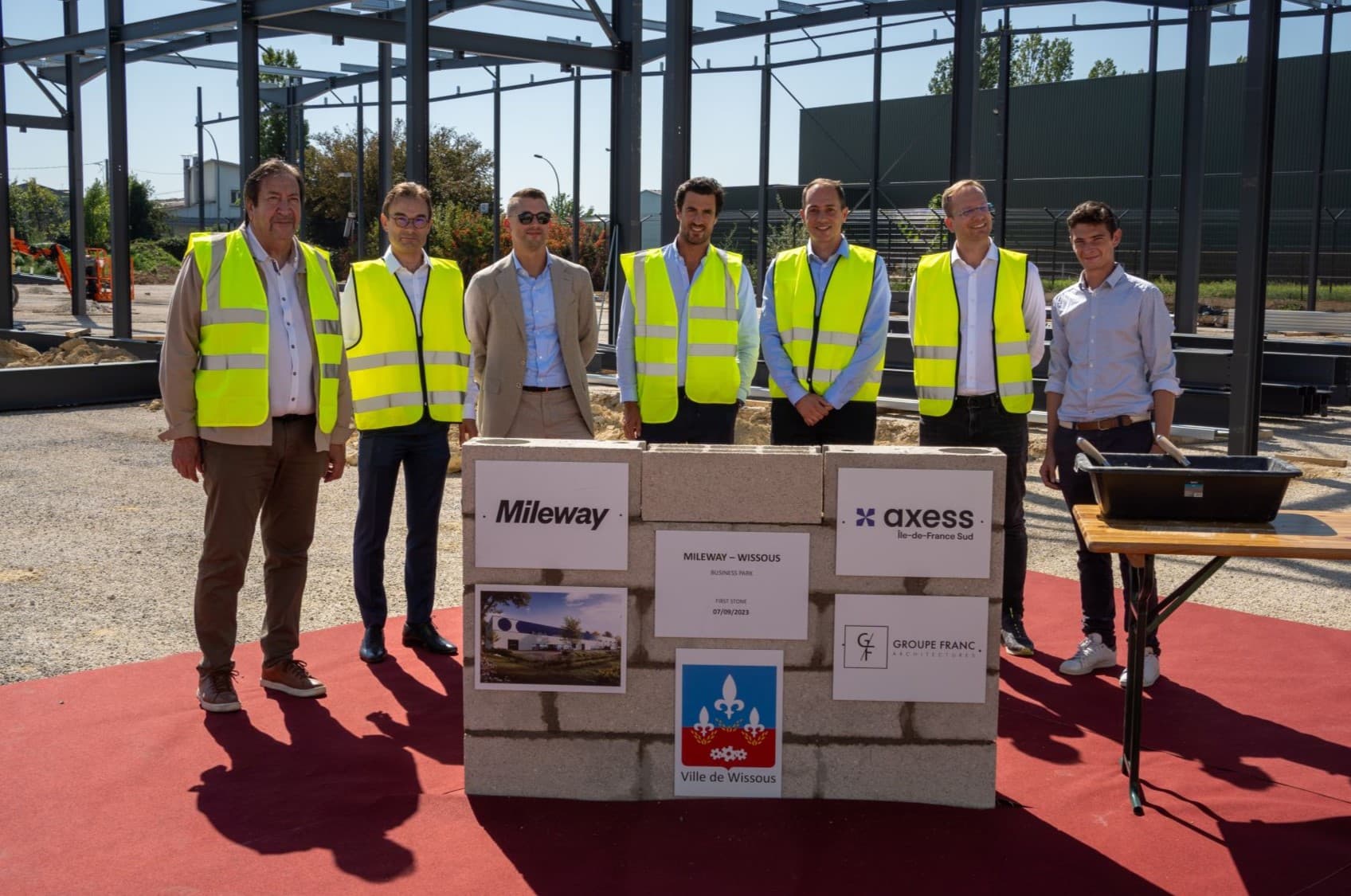 A group of seven people in yellow vests stands behind a construction-themed display with company logos outdoors.