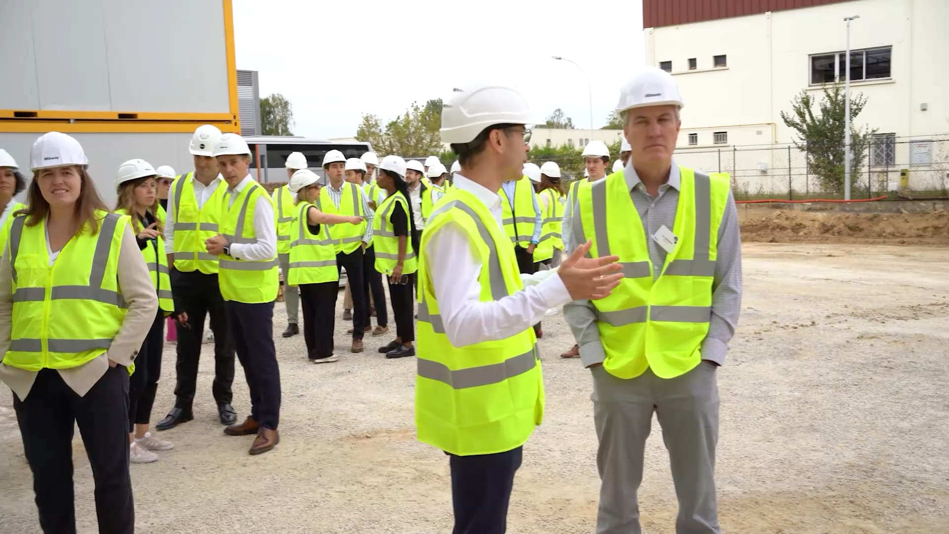 A group of people wearing safety vests and hard hats stand outside, with two individuals talking in the foreground.