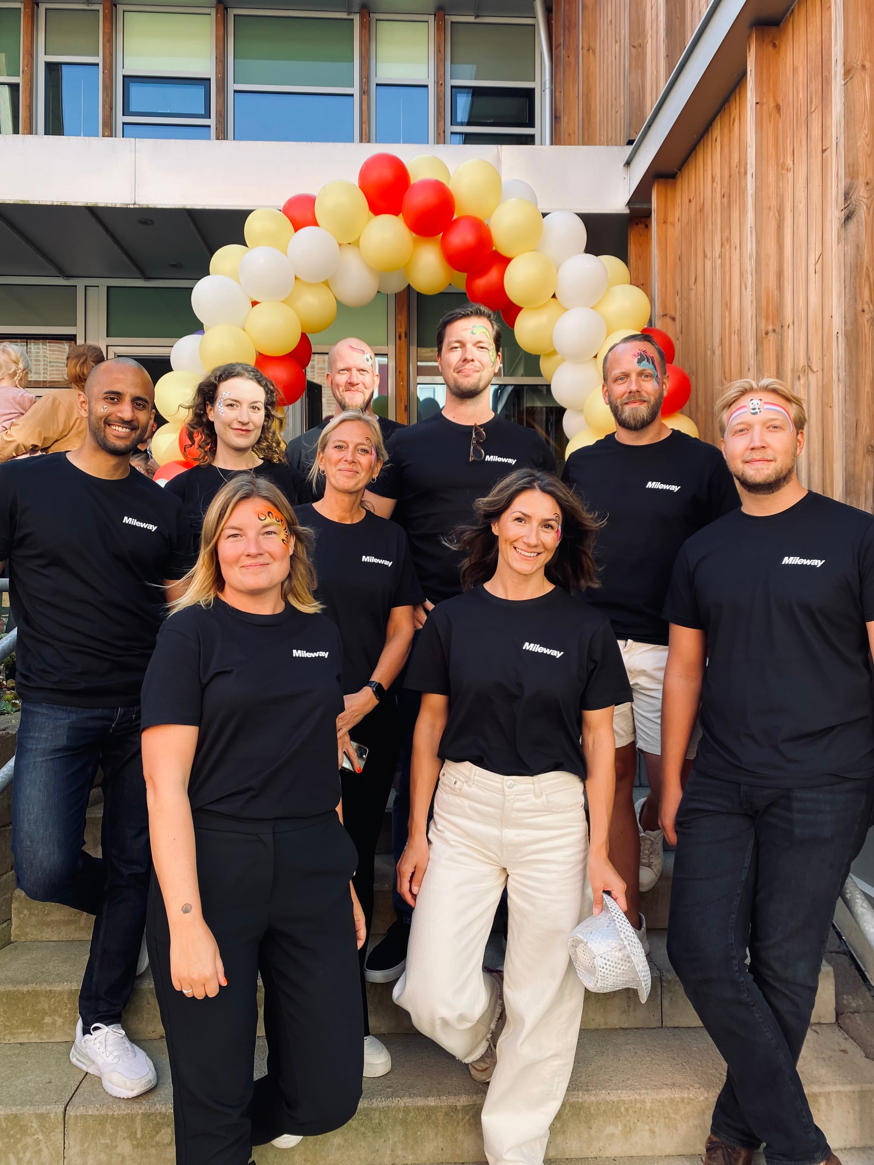 A group of people in black t-shirts stand on steps in front of a red and gold balloon arch.