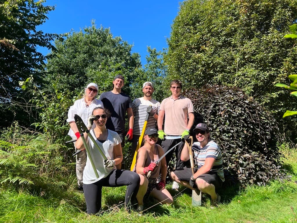 A group of seven people posing outdoors in a garden, holding gardening tools, with trees and a blue sky in the background.