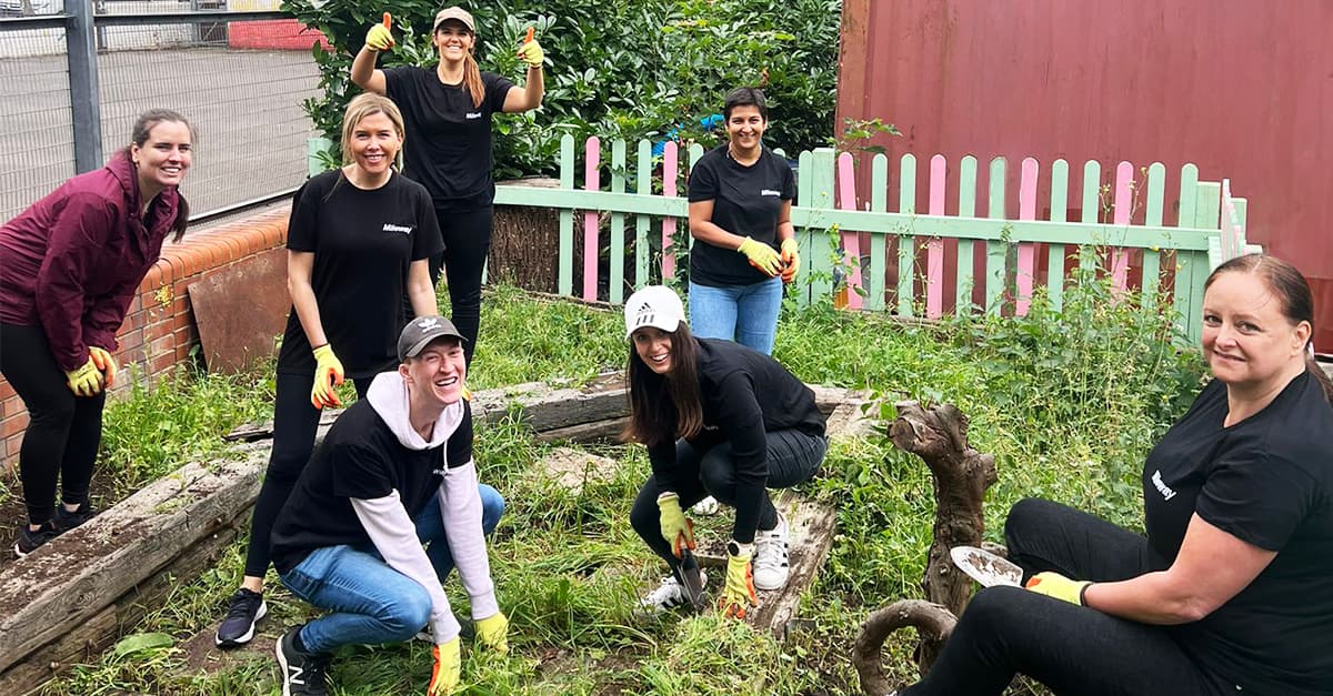 A group of seven people wearing gloves work together in a garden area, surrounded by greenery and a wooden fence. Some are crouching, and others are standing, all appear to be weeding.