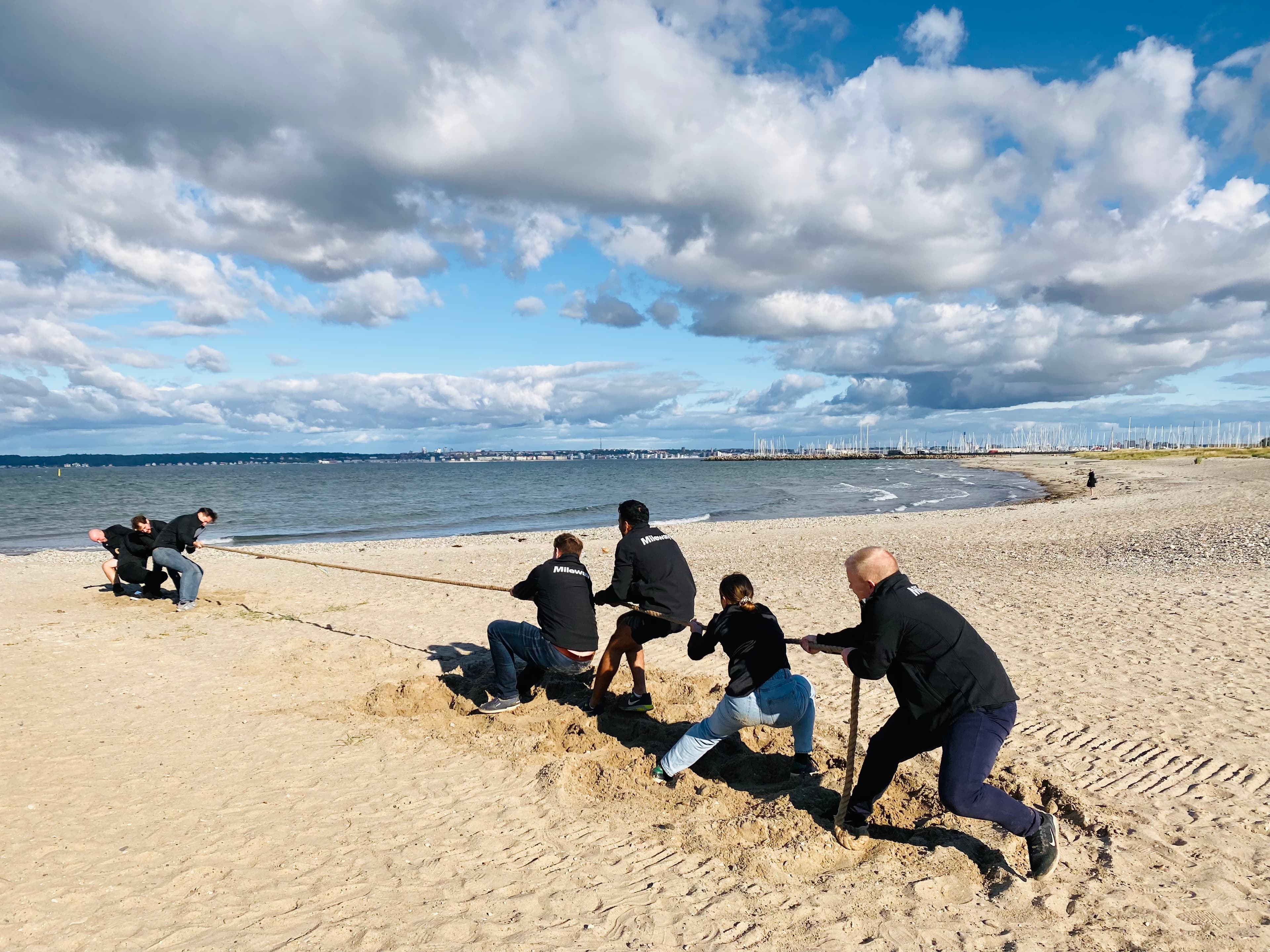 People playing tug-of-war on a sandy beach under a cloudy sky, with the sea and distant shoreline in the background.