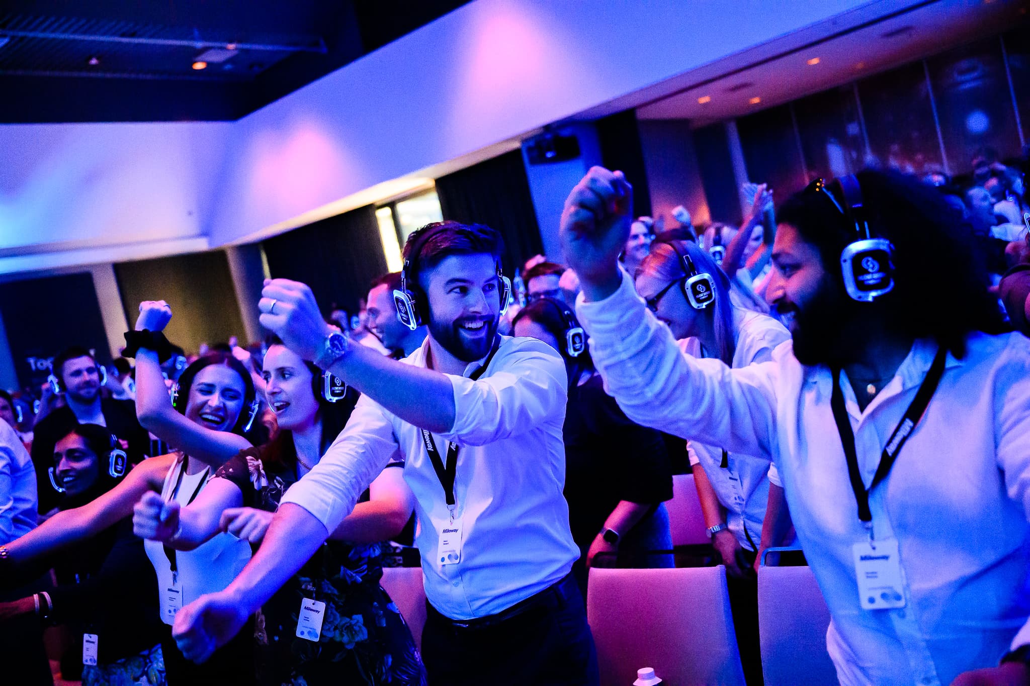 A group of people wearing headphones enjoy a silent disco event indoors, waving their arms and smiling.
