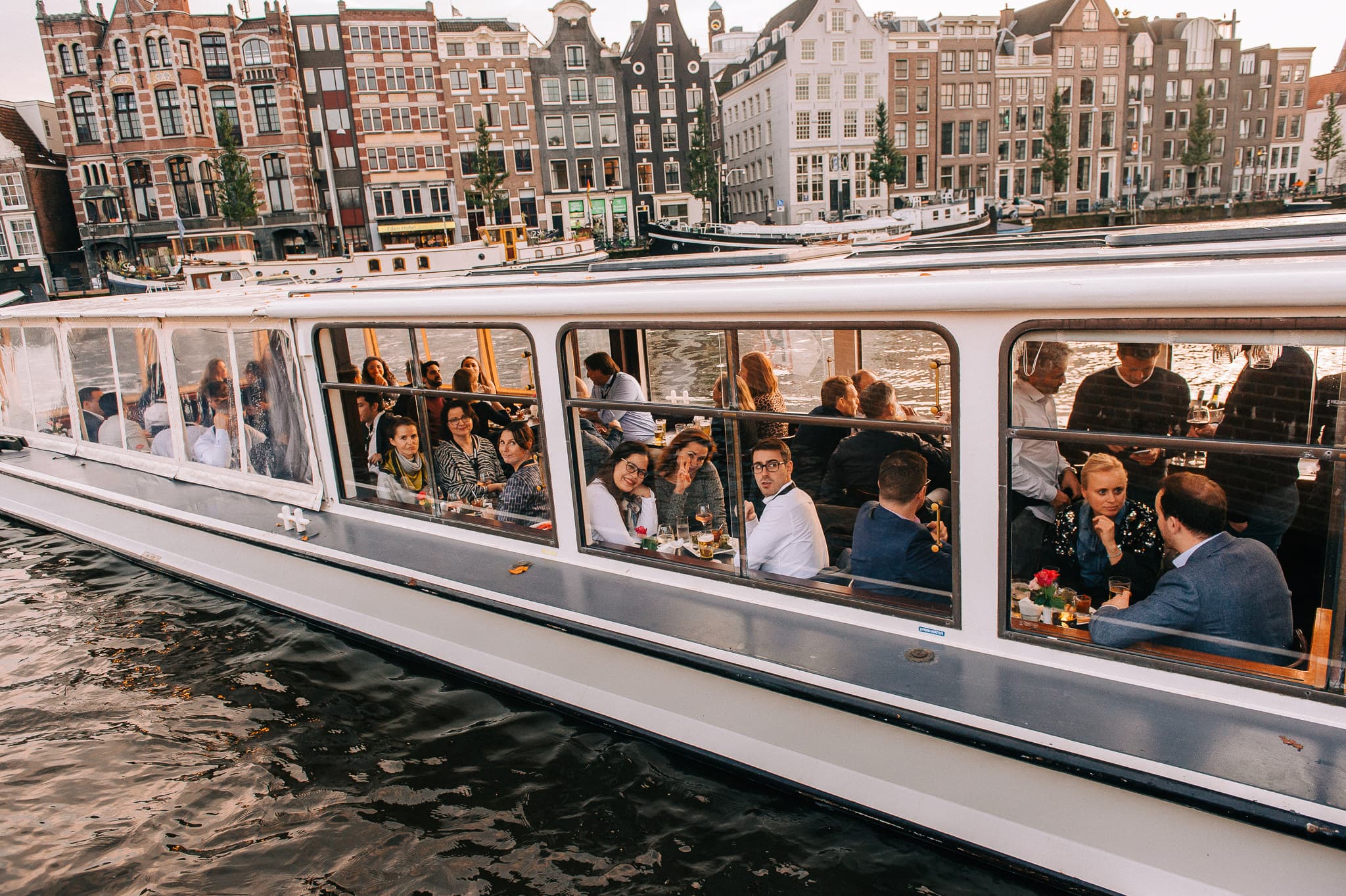 A group of people inside a boat with large windows are enjoying a meal. The boat is on a canal with traditional brick buildings in the background.