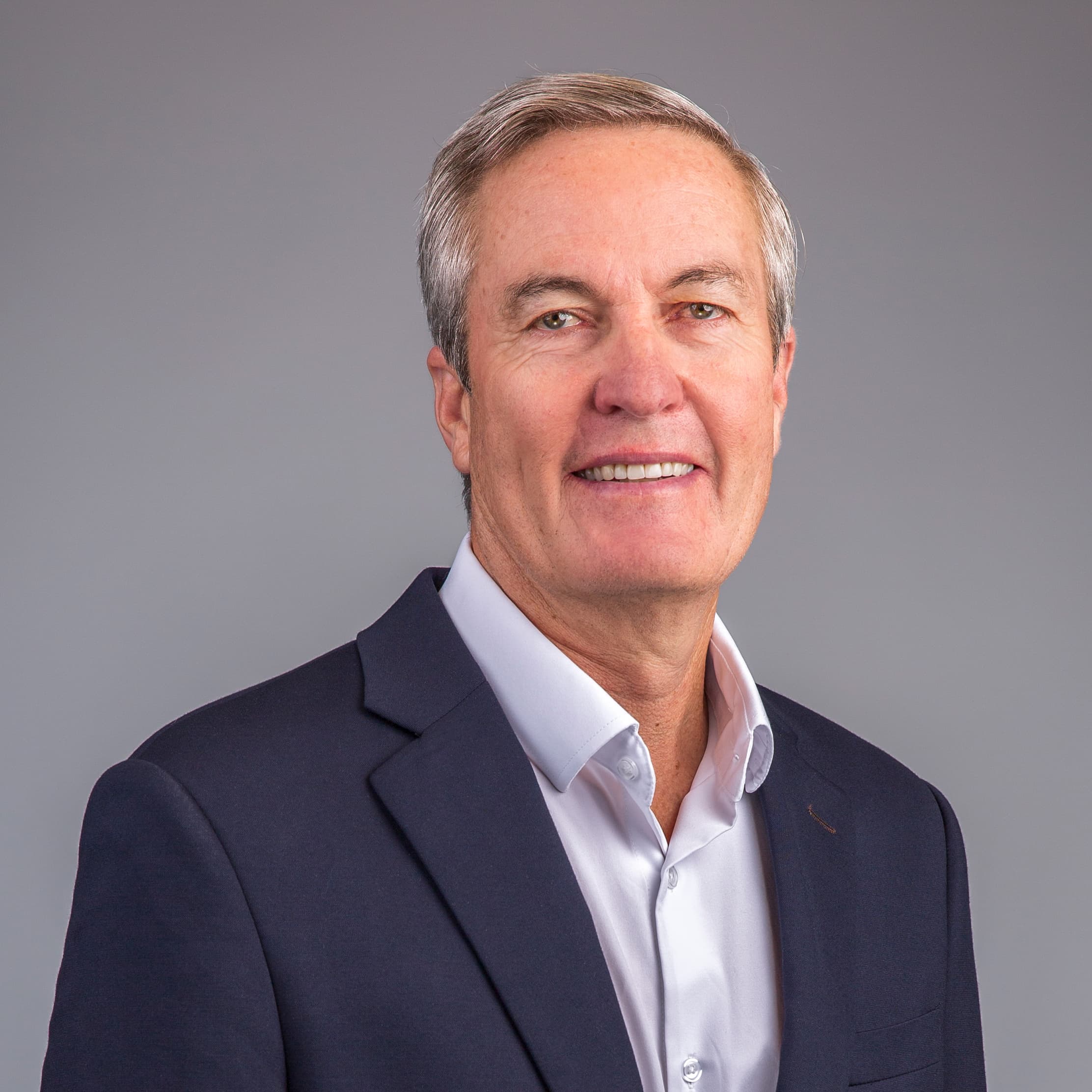 A person wearing a navy blazer and white dress shirt smiles in front of a plain gray background.