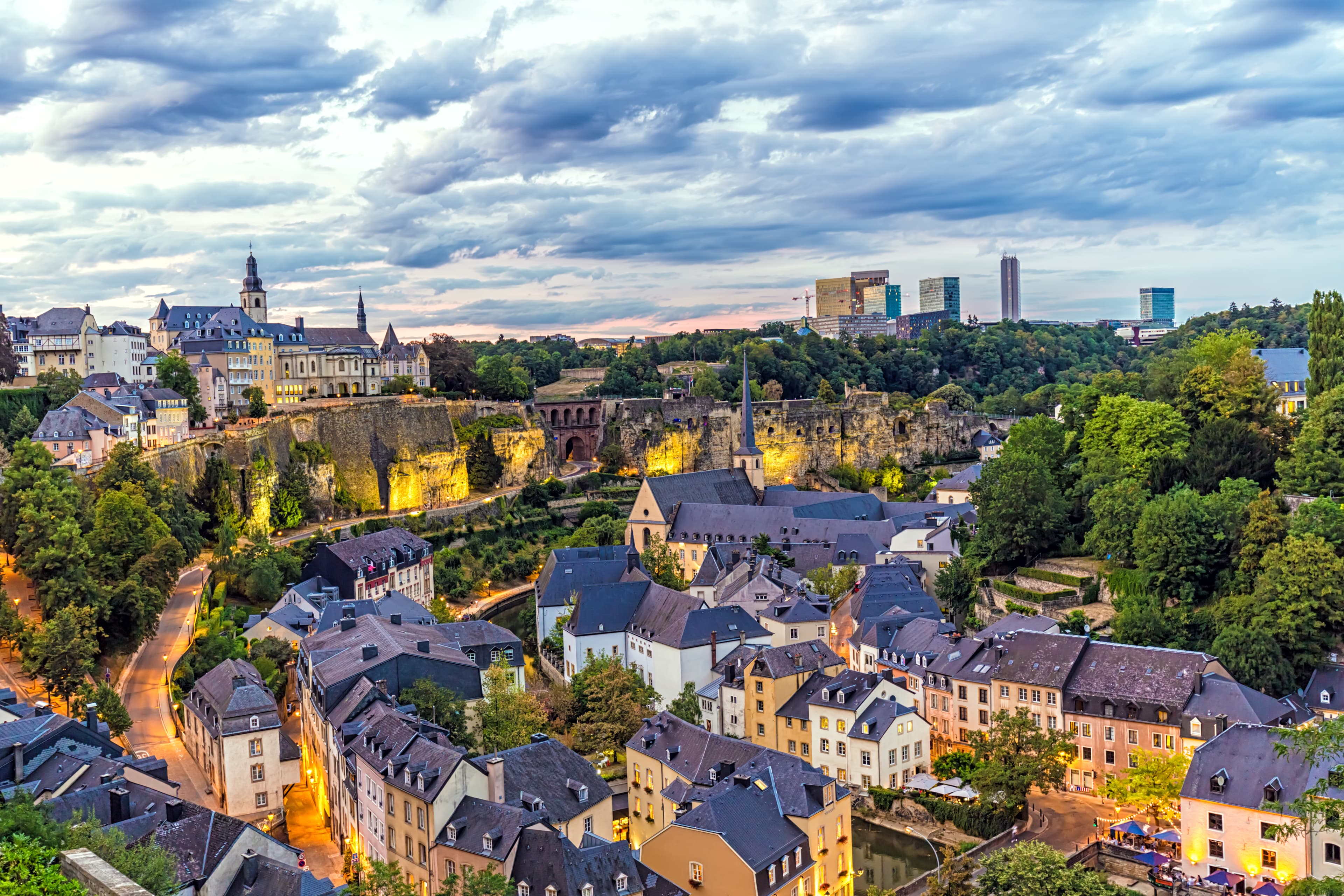 Aerial view of Luxembourg City at sunset, showcasing historic buildings, a church, and greenery under a partly cloudy sky.