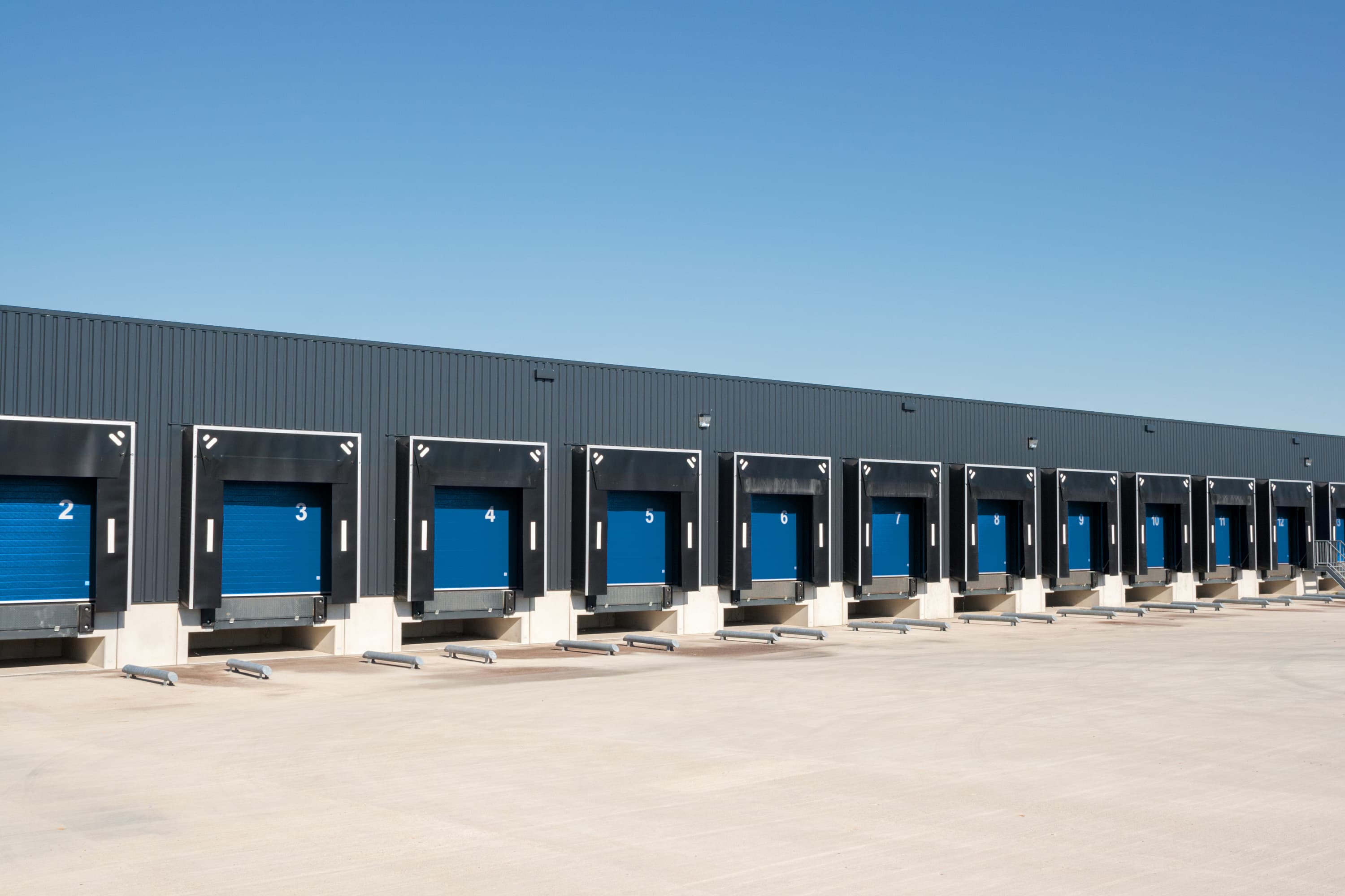 A row of large blue loading dock doors on a warehouse building under a clear blue sky.