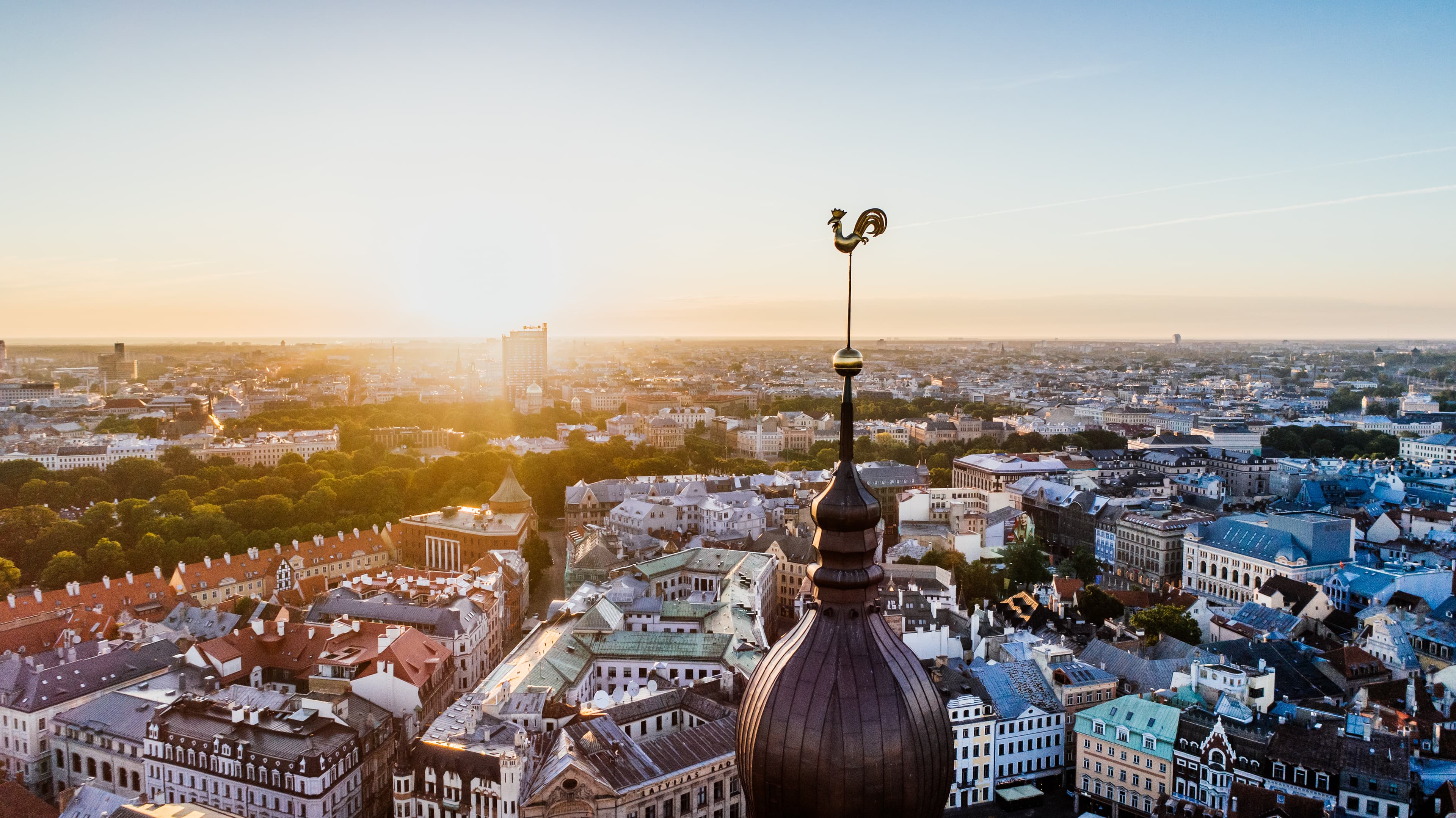 Aerial view of a cityscape at sunset with a prominent church spire featuring a rooster weather vane in the foreground.