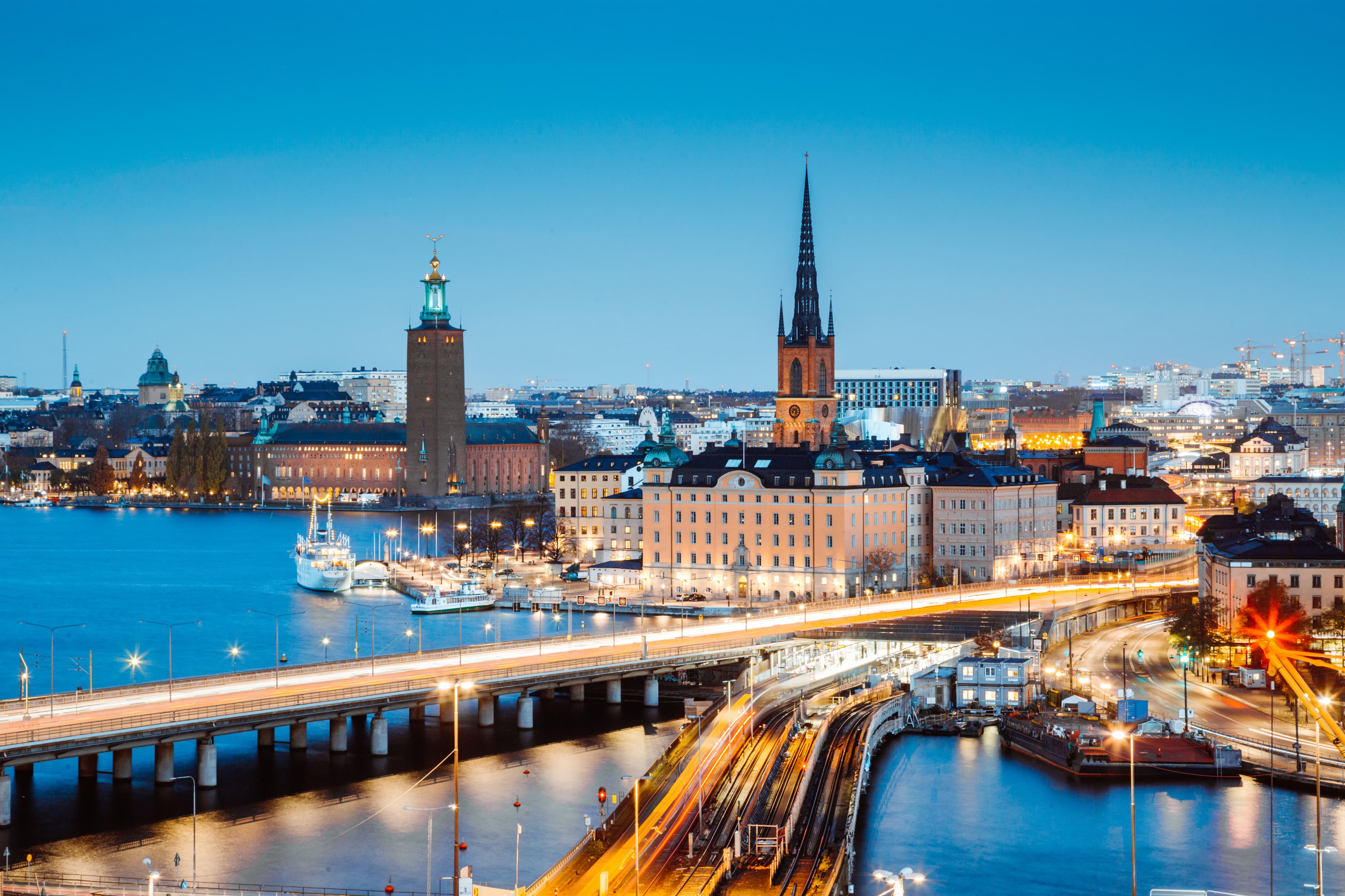 Panoramic view of Stockholm at dusk, featuring illuminated buildings, bridges, and a waterfront with boats.