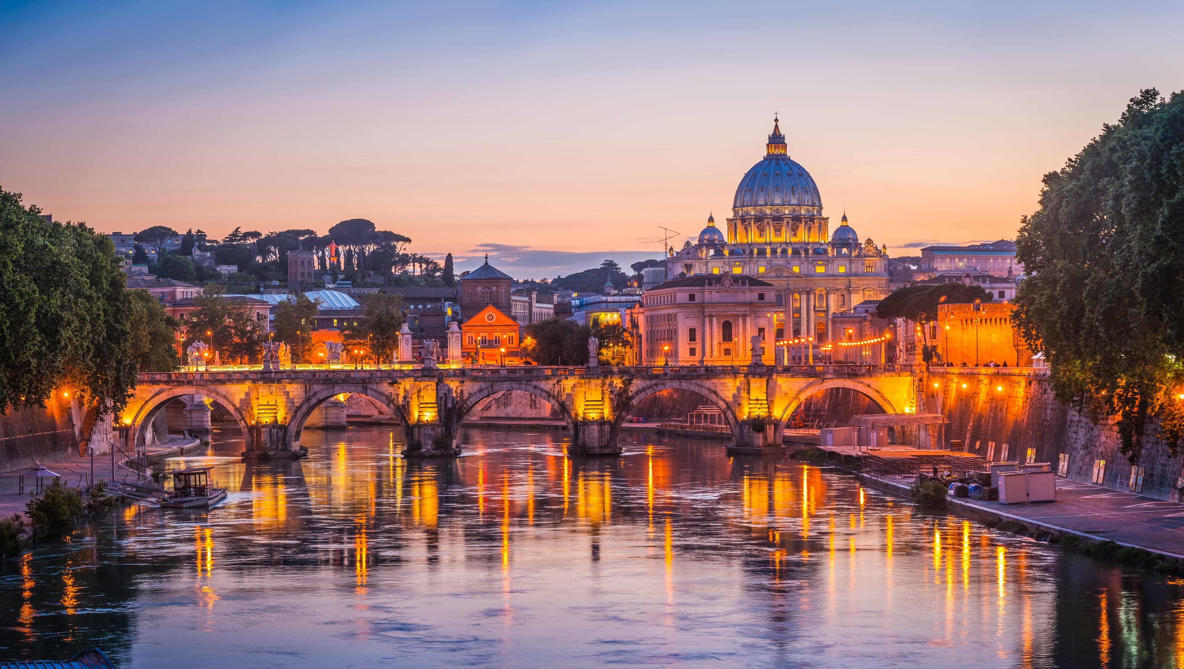 View of St. Peters Basilica at sunset with a bridge over the Tiber River, framed by trees and city lights reflecting on the water.
